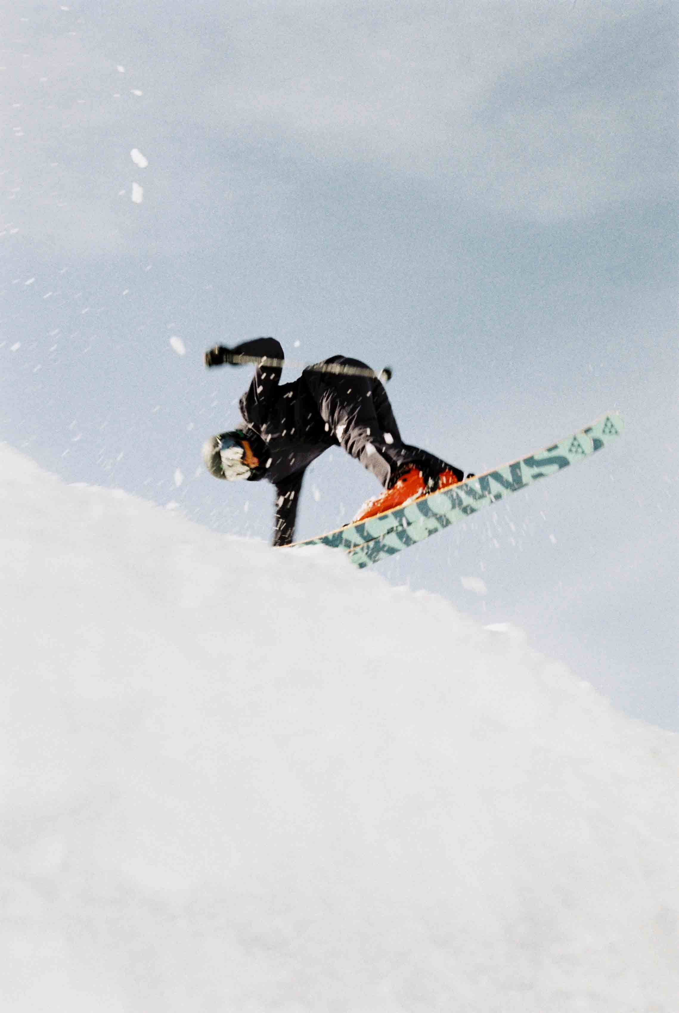 Un skieur en tenue noire et chaussures rouges effectue un saut en snowboard dans la neige sous un ciel clair.