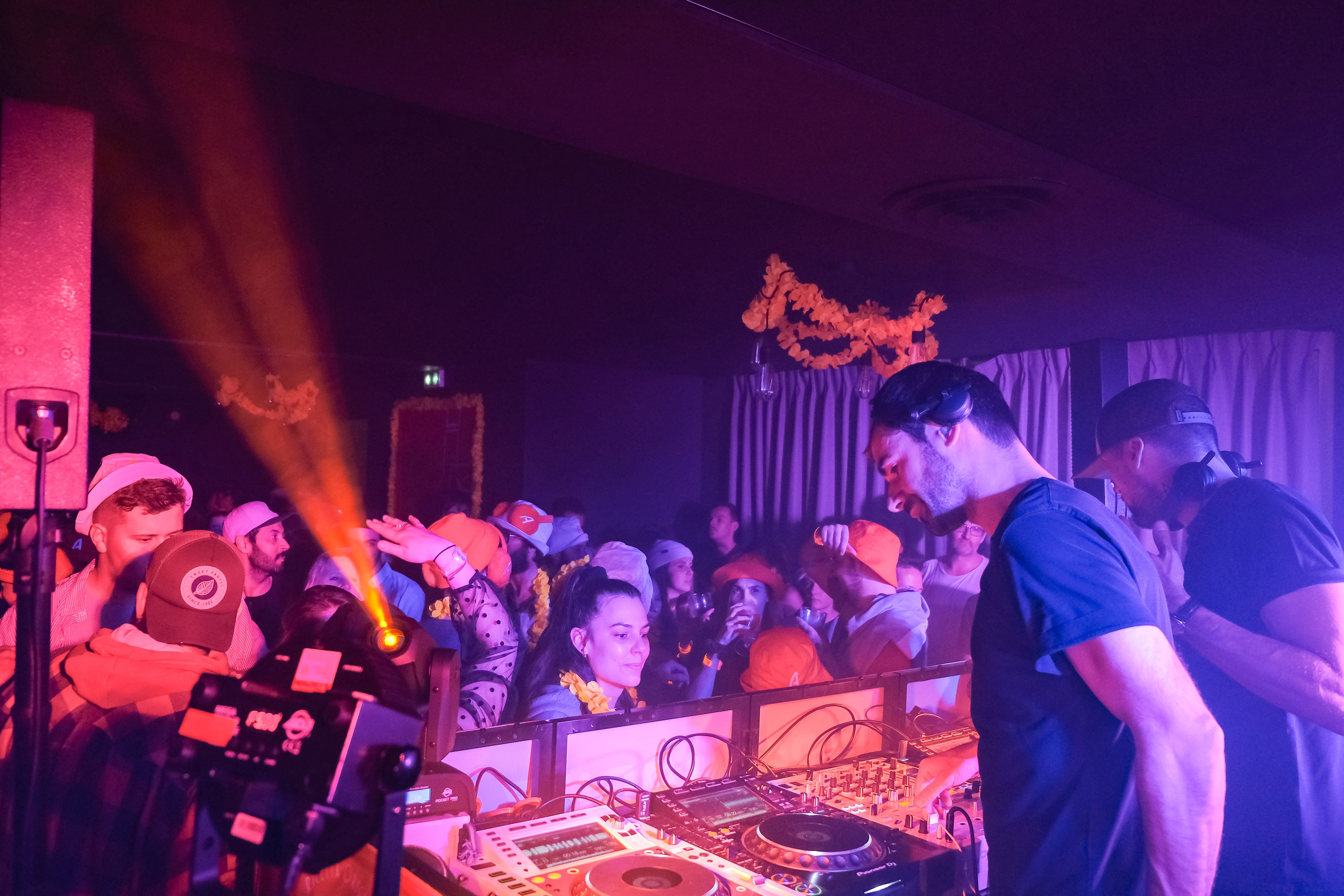 Deux DJs portant des casques, jouant de la musique sur une console de mixage devant une foule animée dans une salle sombre avec des lumières violettes et orange.