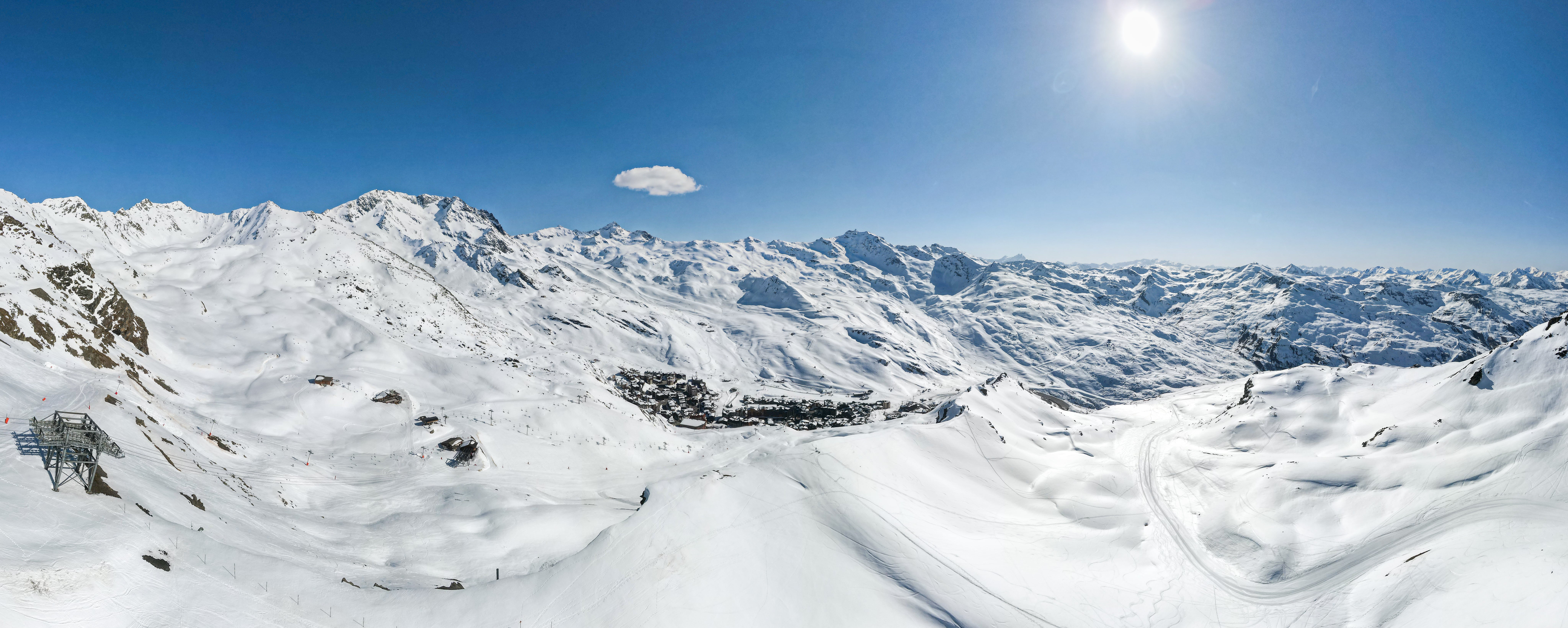 Vue panoramique par temps clair sur une station de ski nichée dans les montagnes enneigées sous un ciel bleu avec un soleil brillant et un petit nuage isolé, incluant des pistes de ski, des installations de remontées mécaniques et des bâtiments au centre.