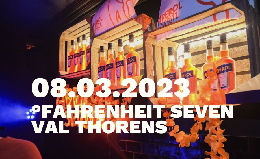 Bouteilles d'Aperol orange illuminées sur des étagères en bois, décorées de guirlandes florales orange, avec un éclairage ambiant bleu en arrière-plan et du texte blanc indiquant la date 08.03.2023 et le lieu Fahrenheit Seven Val Thorens.