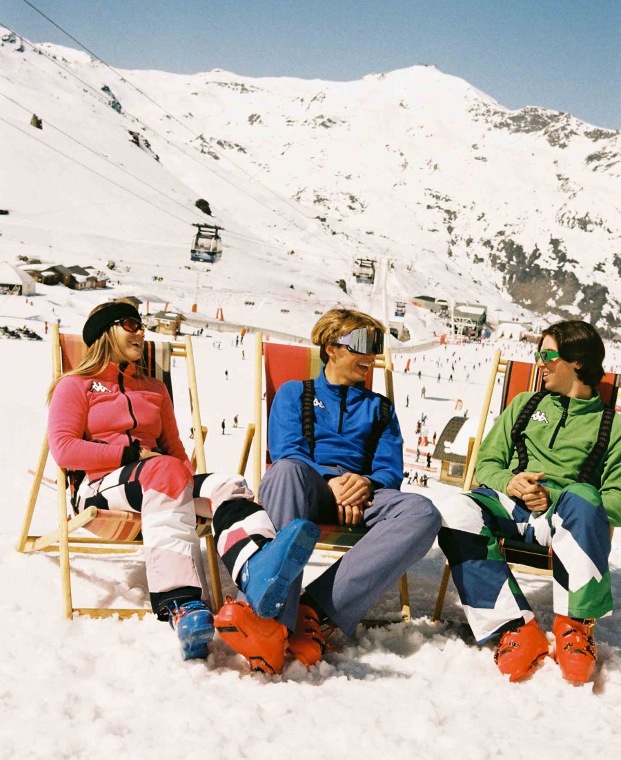 Trois amis en tenue de ski colorée, assis sur des chaises longues dans la neige avec des chaussures de ski, souriant et discutant devant un paysage de montagnes enneigées avec des télécabines et des skieurs en arrière-plan.