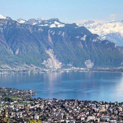 Vue panoramique du lac Léman avec une ville sur la rive avant des montagnes boisées et enneigées en arrière-plan sous un ciel clair.