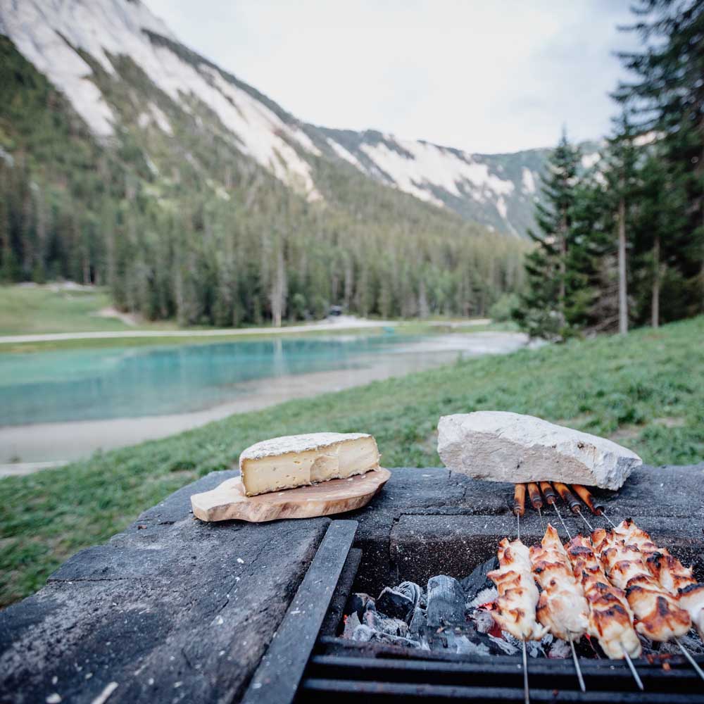 Brochettes de poulet grillées au barbecue en pierre avec un morceau de fromage sur une planche en bois, au bord d'un lac dans une vallée montagneuse verdoyante.