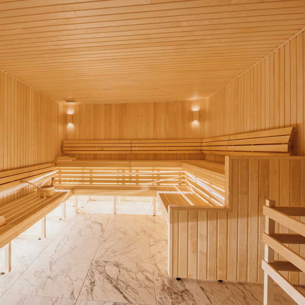 Intérieur d'un sauna avec murs, plafond et bancs en bois clair, sols en marbre blanc, lumière douce aux murs et un seau avec une louche sur un banc.