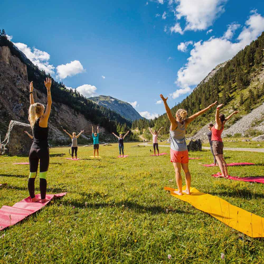 Groupe de personnes en tenue de sport pratiquant le yoga en plein air sur des tapis disposés sur une prairie entourée de montagnes sous un ciel bleu partiellement nuageux.