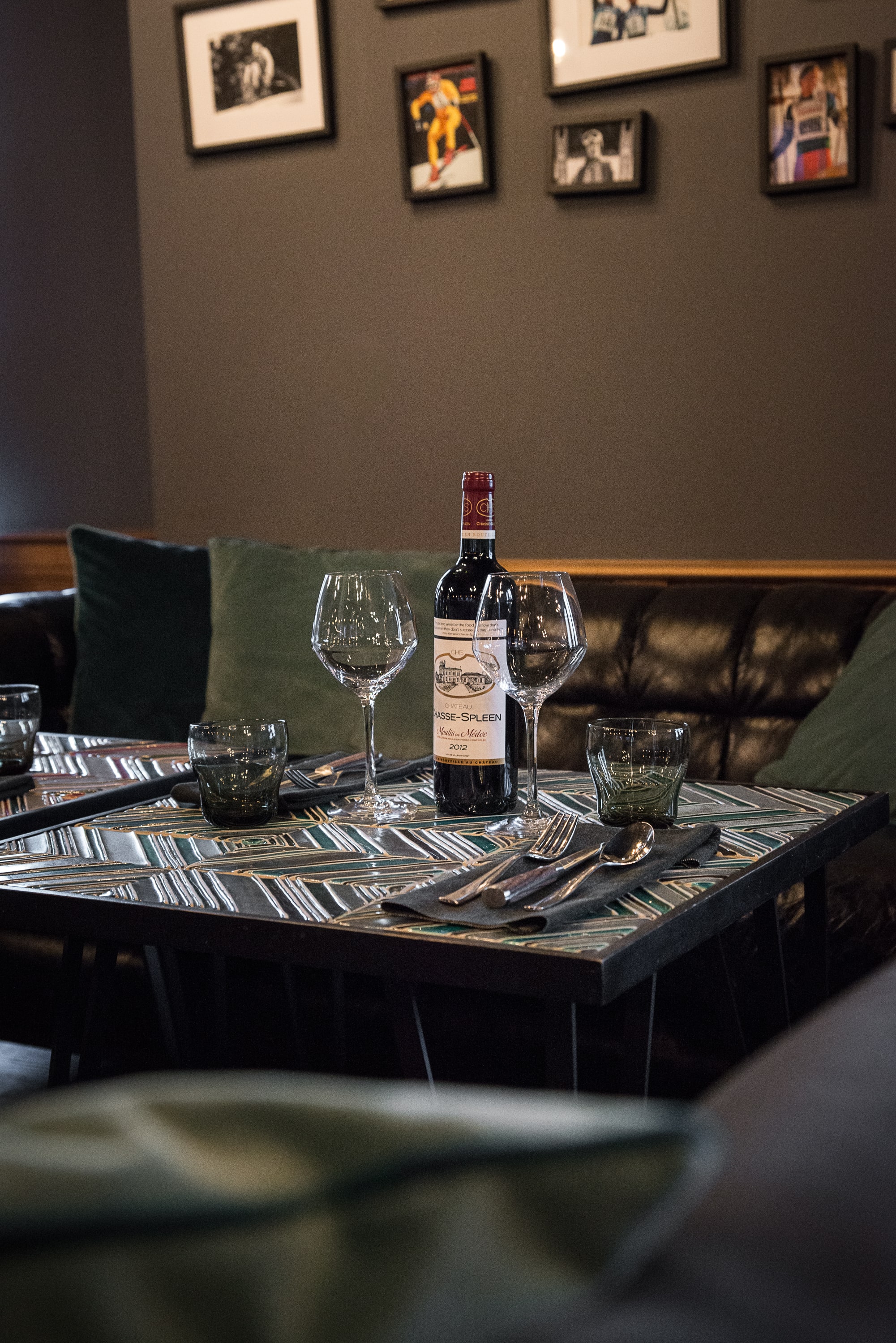 Bouteille de vin rouge Château Lespéeen 2012 et deux verres à vin sur une table décorée, avec des couverts sur une serviette noire, dans un coin salon avec des coussins verts et un canapé en cuir noir, mur gris avec des cadres photo.