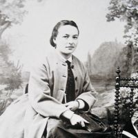 Portrait en noir et blanc d'une femme assise, habillée d'une veste claire sur une robe sombre, avec des cheveux lisses tirés en arrière, posant devant un décor naturel flou.
