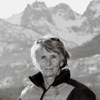 Portrait en noir et blanc d'une femme âgée portant une veste polaire et des lunettes de soleil sur la tête, avec des montagnes enneigées en arrière-plan.