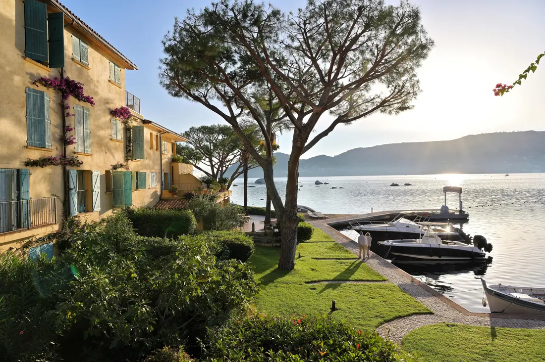 Vue sur une pelouse verte et un jardin avec des arbres sous un ciel clair, un bâtiment jaune à volets bleus sur la gauche, et un quai avec plusieurs bateaux amarrés au bord d'une étendue d'eau calme au soleil couchant, avec des montagnes en arrière-plan.