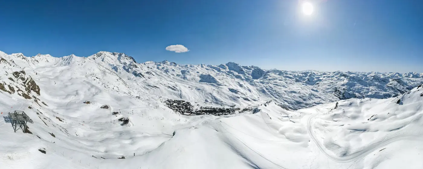 Panorama ensoleillé d'une station de ski entourée de montagnes enneigées sous un ciel bleu avec un petit nuage blanc.