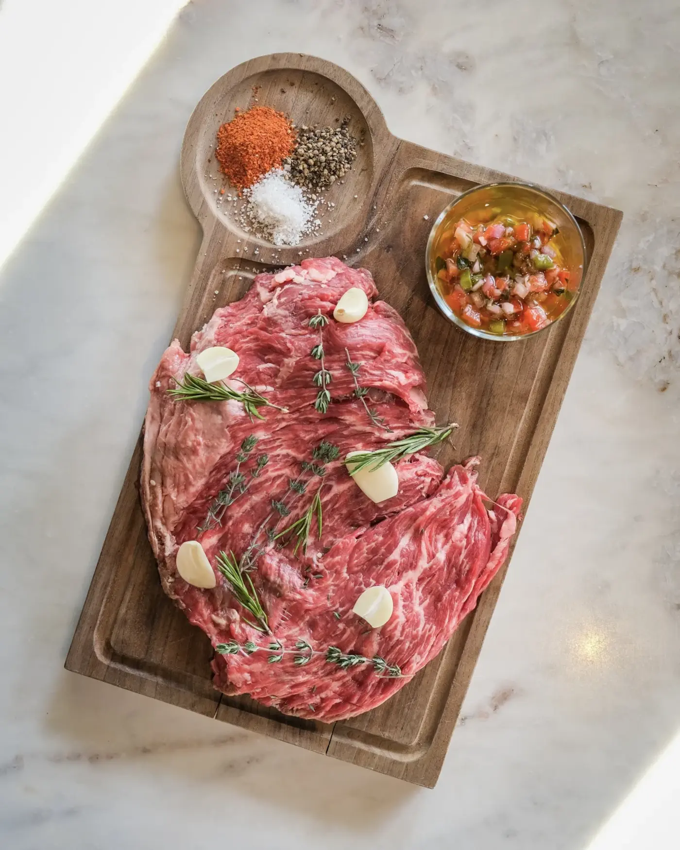 Une planche en bois avec une grande pièce de viande crue garnie de gousses d'ail et de branches de romarin et de thym, accompagnée d'un petit bol de sauce salsa et de trois piles d'épices : paprika, poivre noir et sel.