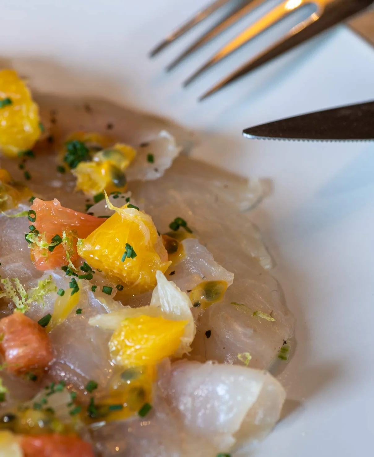 Carpaccio de poisson blanc sur assiette blanche, garni de petits morceaux d'agrumes, de ciboulette ciselée et de graines de fruit de la passion, avec une fourchette et un couteau en arrière-plan.