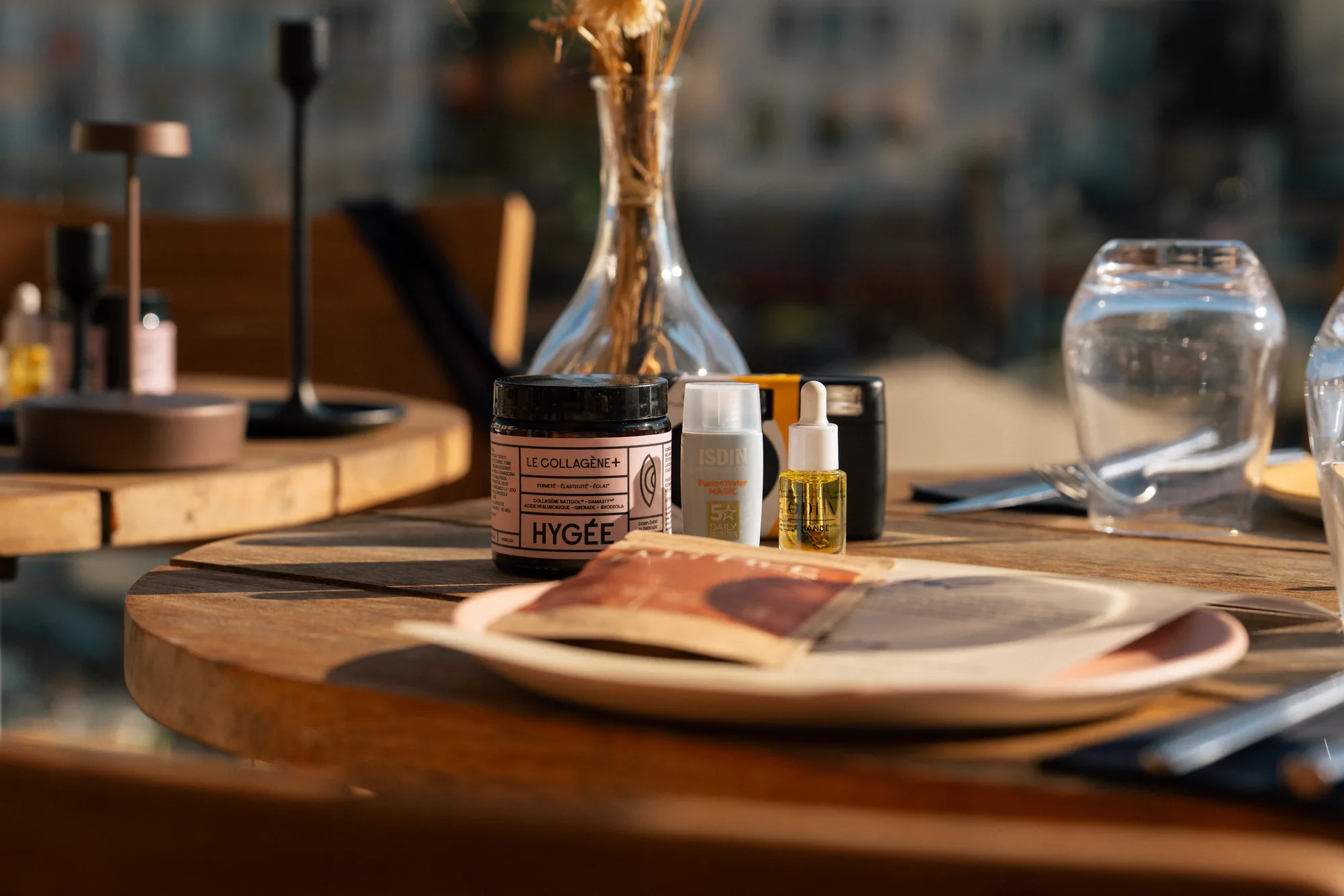 Pot de complément alimentaire Le Collagène+ Hygée, flacon ISDIN FusionWater Magic, sérum avec compte-gouttes, verre d'eau et assiette sur une table en bois dans un cadre lumineux.