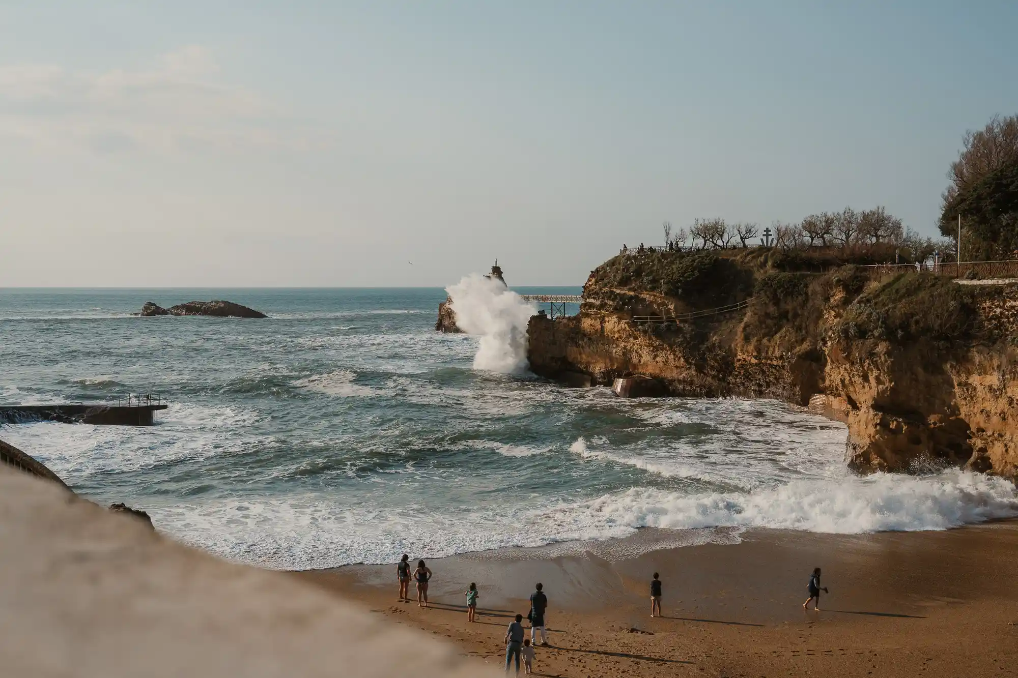 Vue sur une plage avec plusieurs personnes debout sur le sable regardant les vagues qui éclatent contre des falaises rocheuses sous un ciel clair.