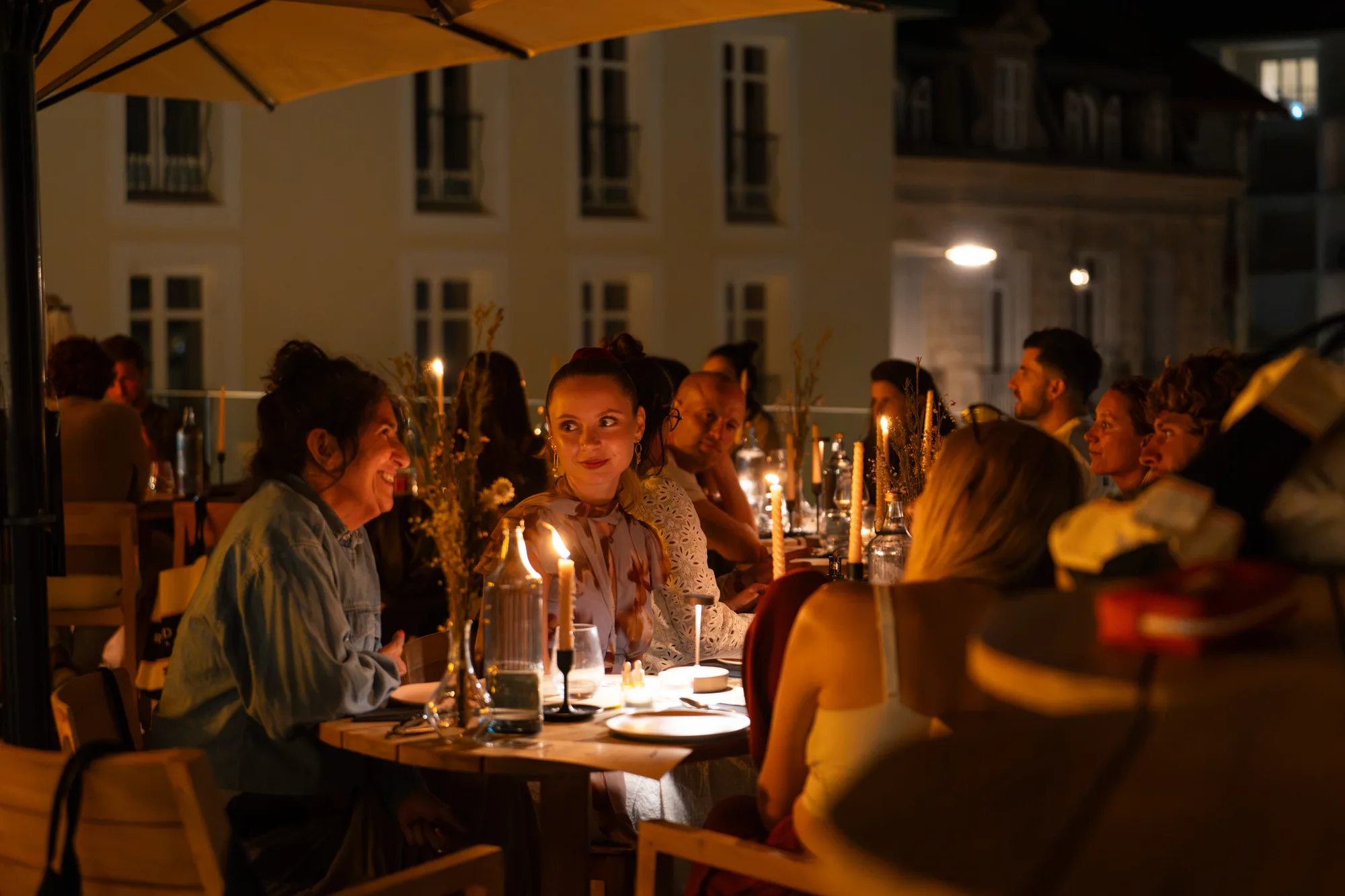 Groupe de personnes assises autour d'une table en plein air le soir, éclairées par des bougies allumées, sous un grand parasol jaune, dans une ambiance chaleureuse et conviviale.