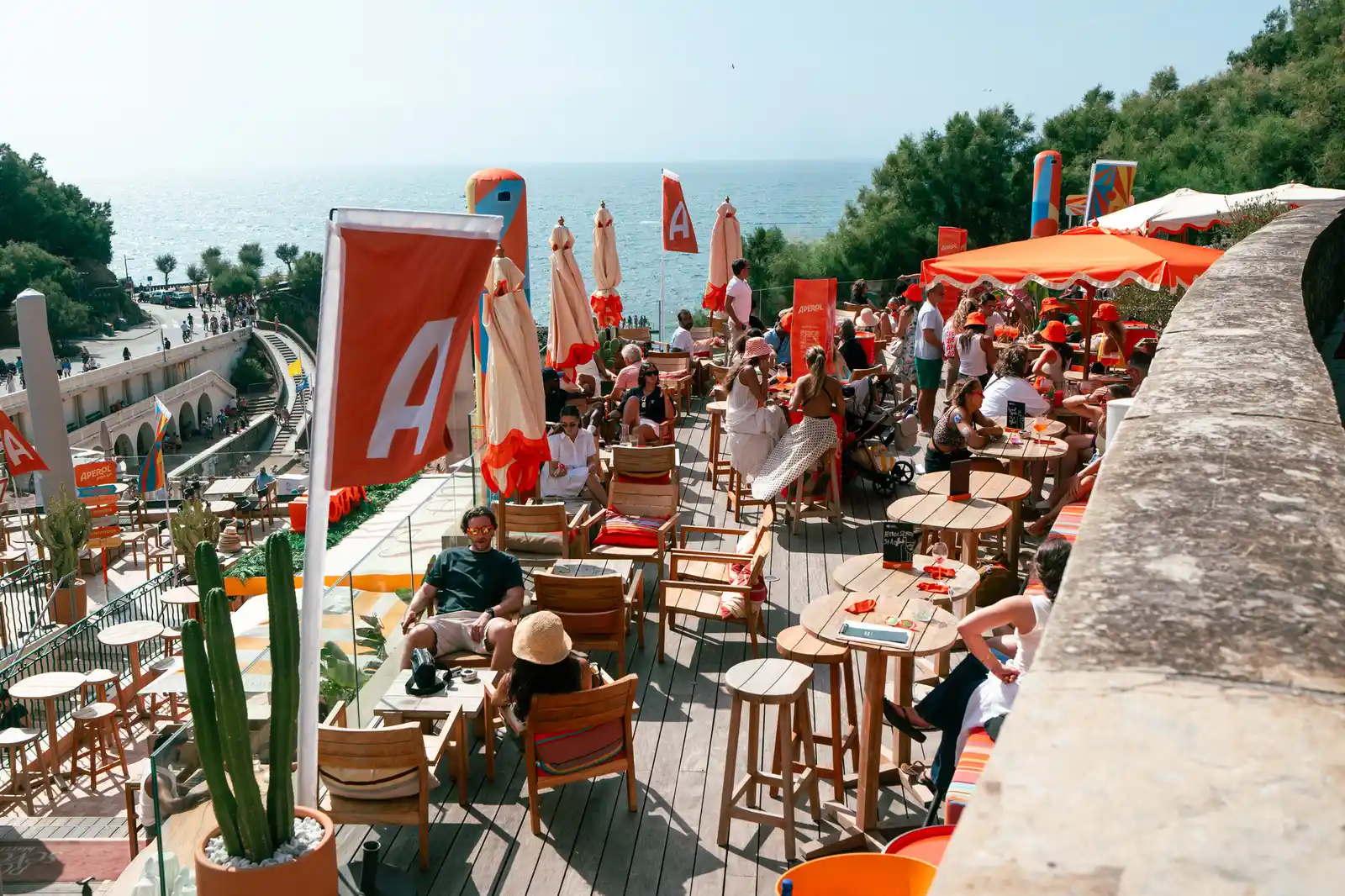 Terrasse ensoleillée d'un bar en bord de mer avec des parasols orange clair, des tables en bois, des chaises, des clients assis discutant et profitant de boissons, vue sur la mer et une promenade animée à gauche.