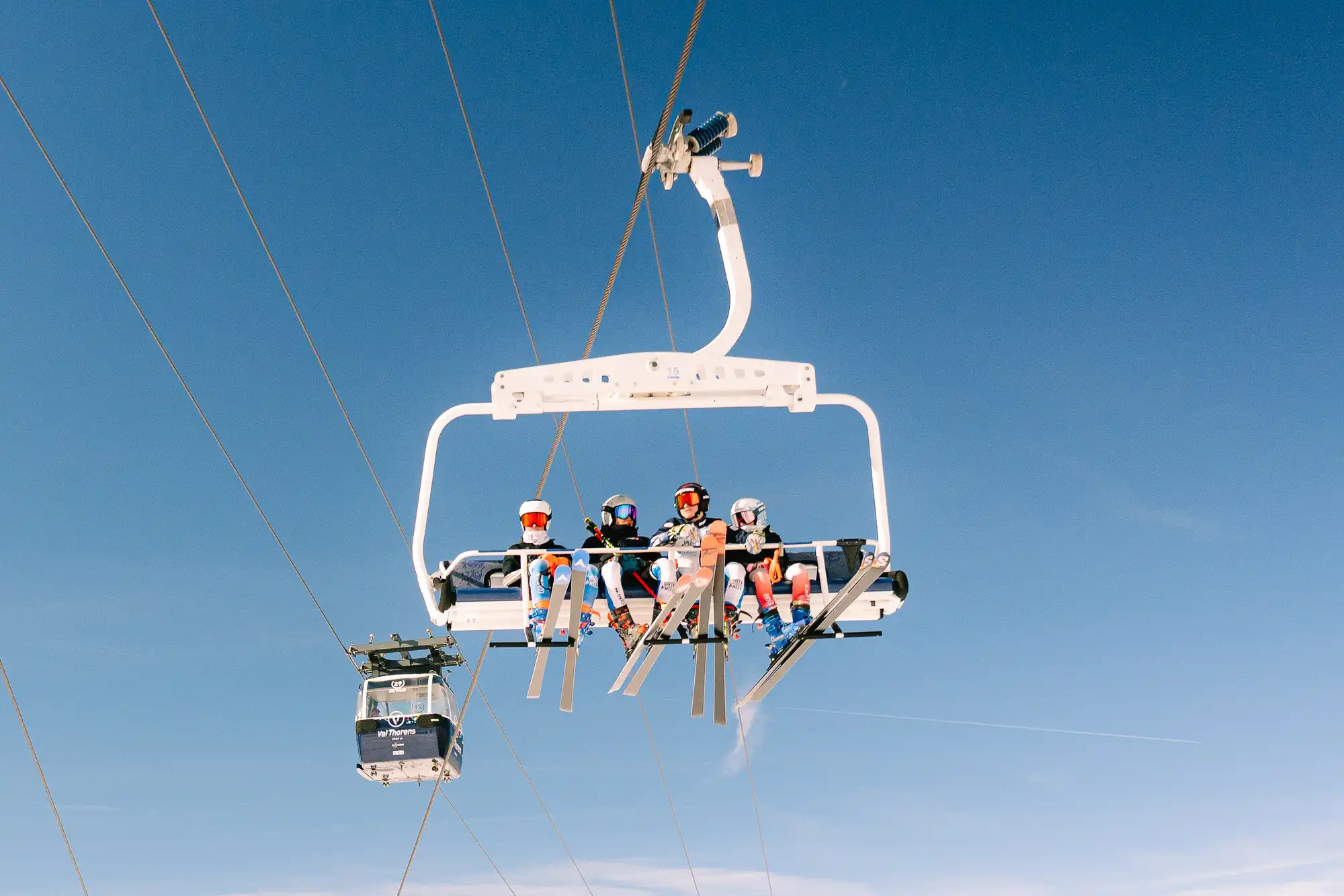 Quatre skieurs en combinaison et casque blanc montent sur un télésiège blanc suspendu dans le ciel bleu clair, avec une cabine de télécabine en arrière-plan.