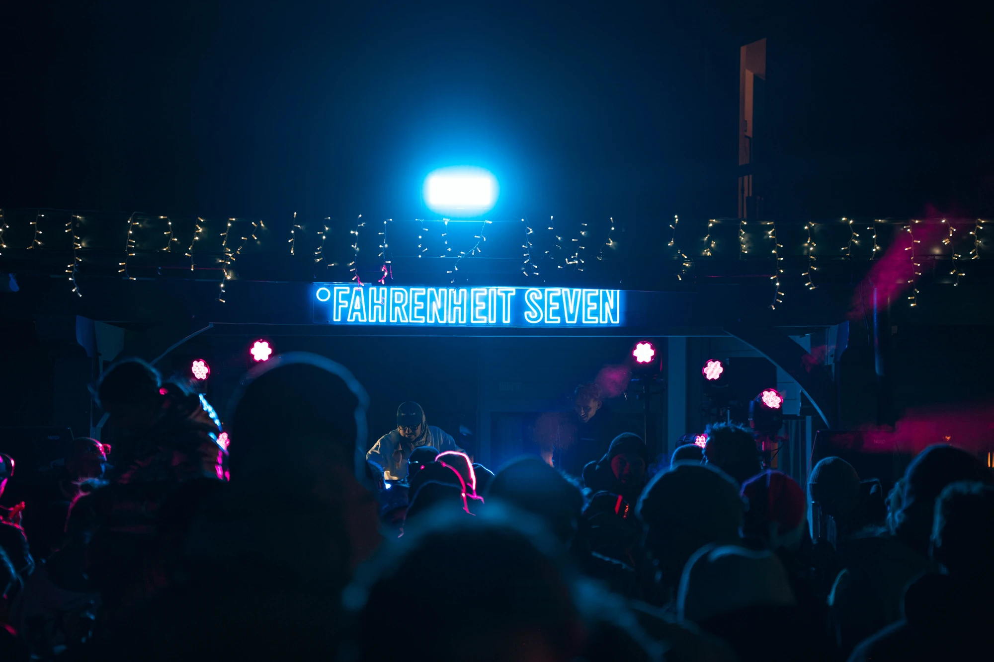 Foule devant une scène extérieure de nuit avec un panneau lumineux bleu indiquant "FAHRENHEIT SEVEN" et des guirlandes lumineuses décoratives au-dessus.