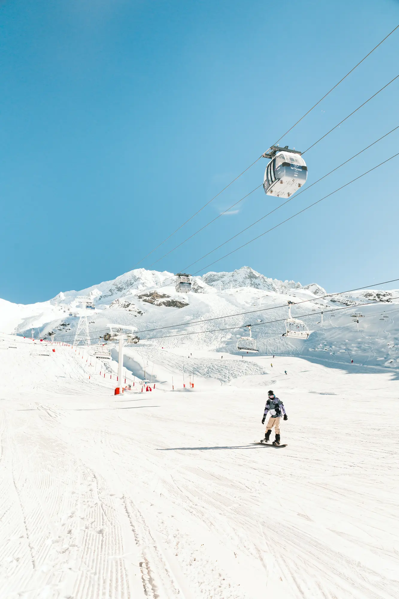 Un snowboarder descend une piste enneigée dans une station de ski ensoleillée avec des remontées mécaniques et des télécabines visibles, sous un ciel bleu clair et des montagnes enneigées en arrière-plan.
