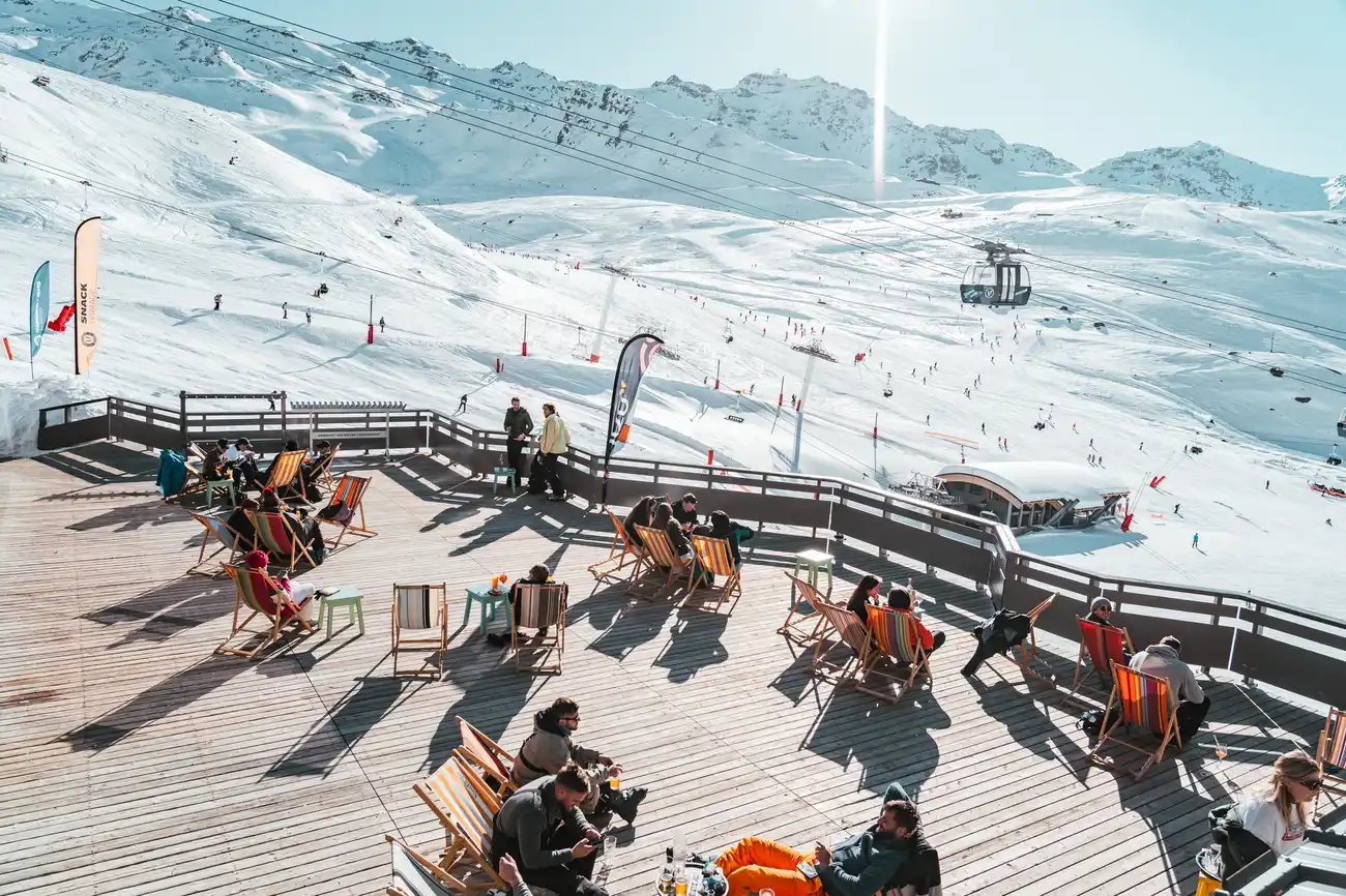 Terrasse en bois avec des personnes assises sur des chaises longues colorées profitant du soleil, avec en arrière-plan des montagnes enneigées et des skieurs sur les pistes, ainsi qu'une télécabine suspendue au-dessus des pistes.