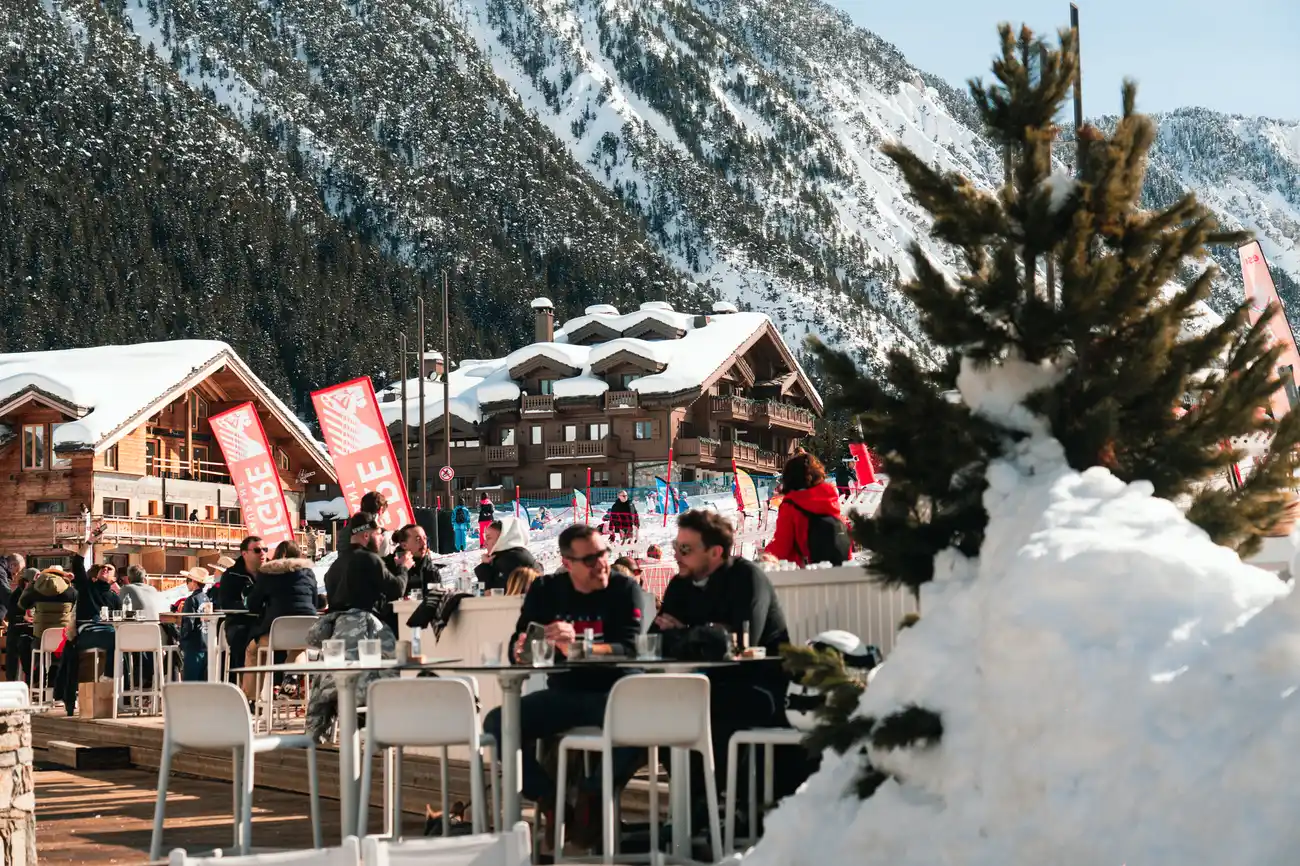 Des personnes profitent d'une terrasse ensoleillée avec des tables et des chaises devant des bâtiments en bois enneigés, avec des montagnes boisées et enneigées en arrière-plan, un petit sapin recouvert de neige au premier plan droite.
