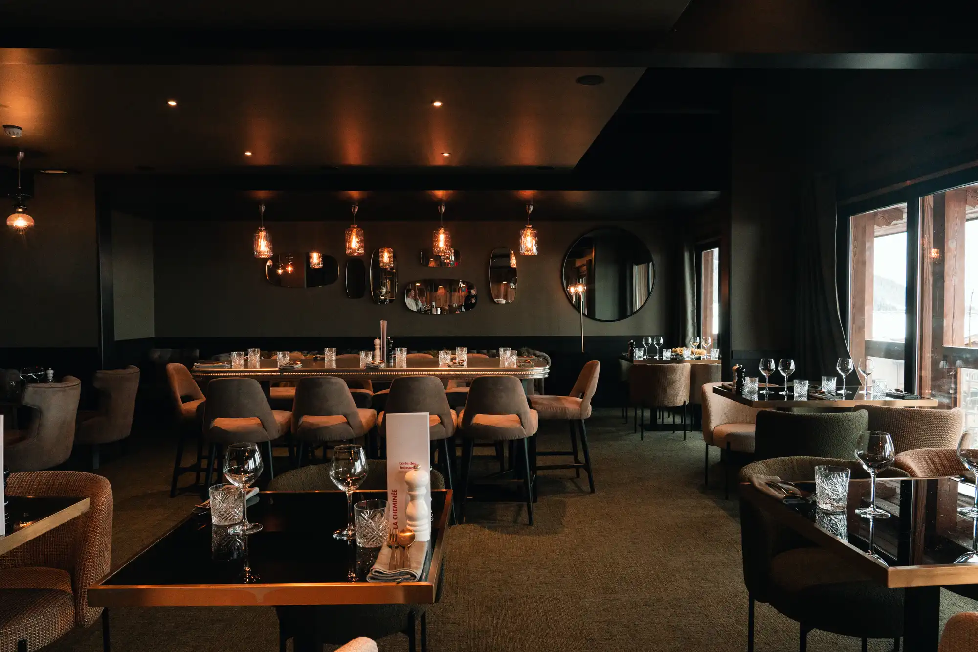 Intérieur d'un restaurant élégant avec des tables dressées, des chaises confortables, un éclairage tamisé par des suspensions en verre et des miroirs décoratifs sur un mur sombre, et une vue sur l'extérieur à travers de grandes fenêtres.