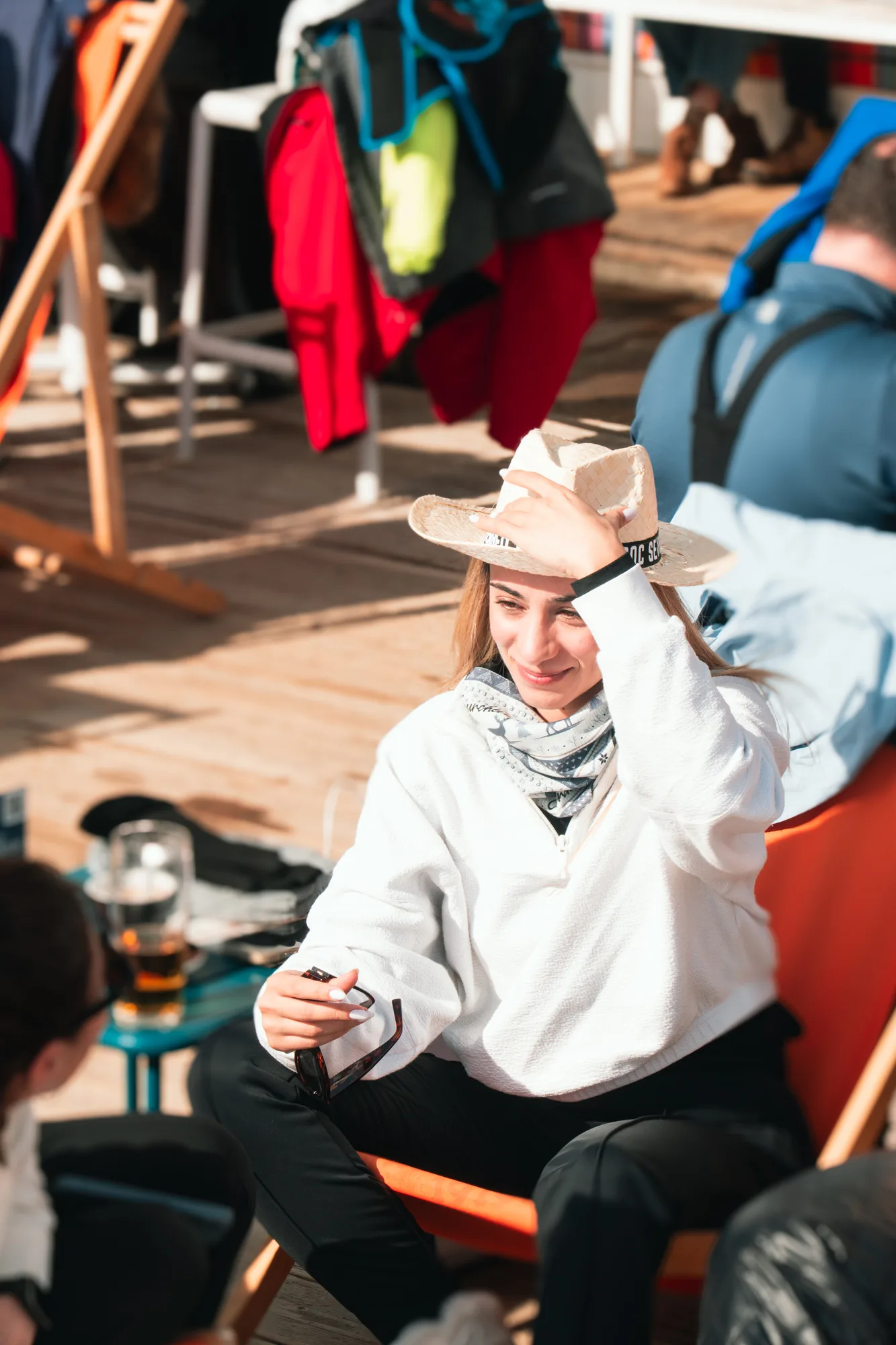 Une femme assise sur une chaise orange en bois, portant un chapeau de paille, un sweat blanc et un foulard autour du cou, souriante et tenant une paire de lunettes noires, en conversation avec une autre personne dans un environnement lumineux avec d'autres personnes en arrière-plan.
