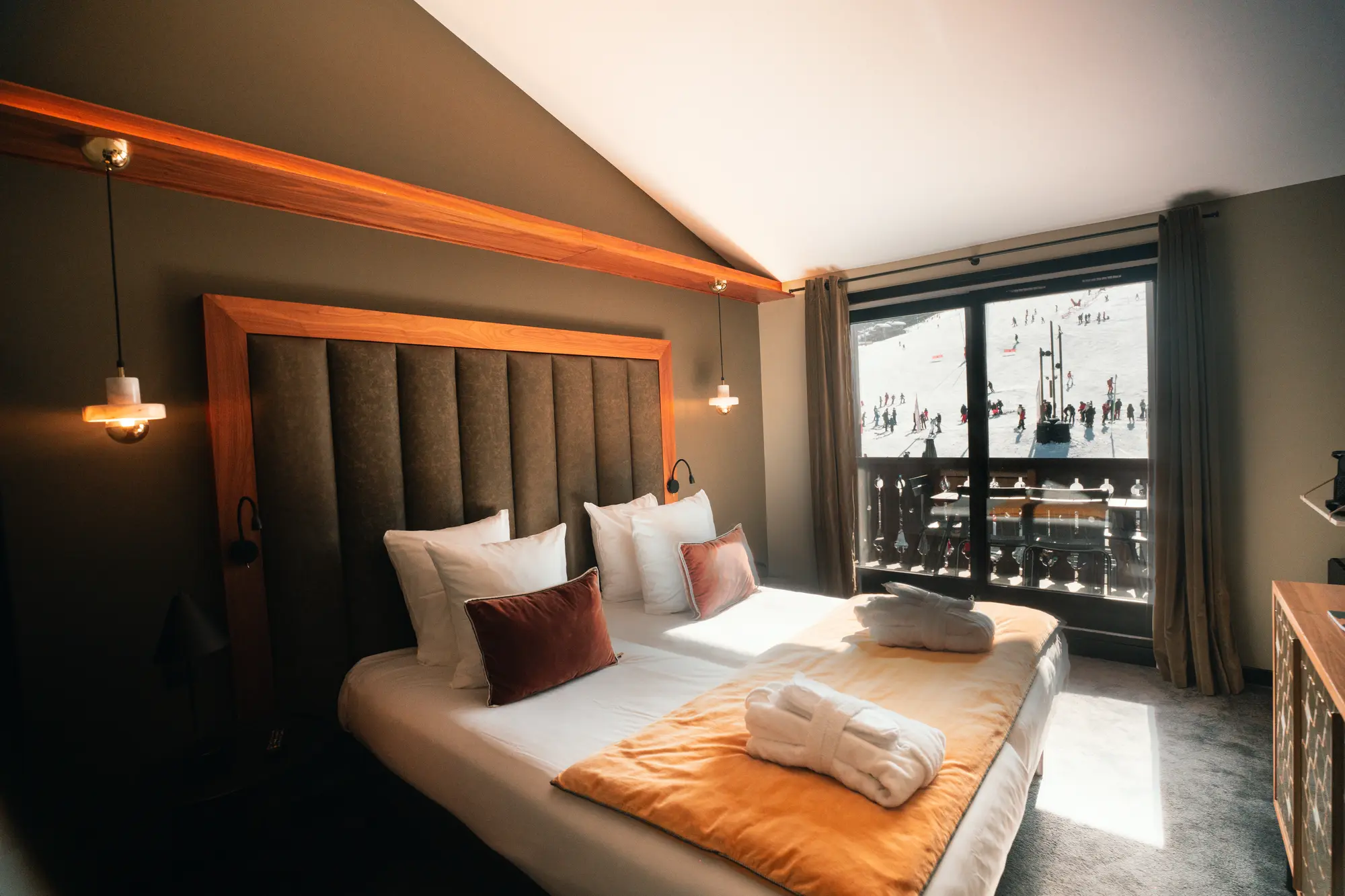 Chambre d'hôtel avec un lit double fait, coussins blancs et marron, deux peignoirs blancs sur une couverture jaune, et une grande fenêtre donnant sur une scène de ski animée en extérieur.