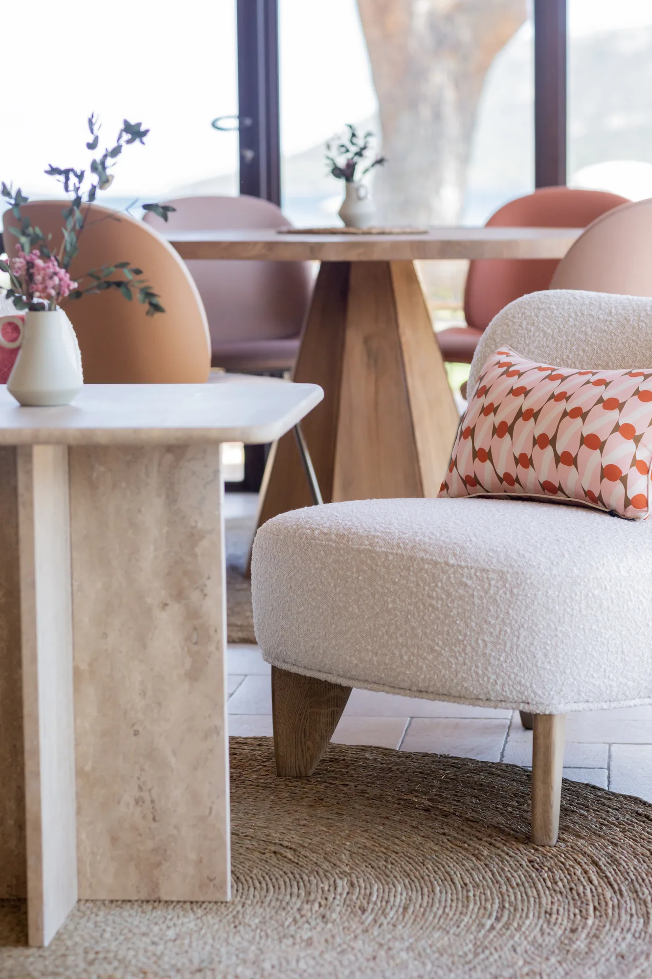 Coin salon avec fauteuil blanc texturé sur tapis tressé, coussin décoratif à motifs géométriques orange et rose, table en pierre avec vase à fleurs, et table en bois entourée de chaises roses devant une grande fenêtre.