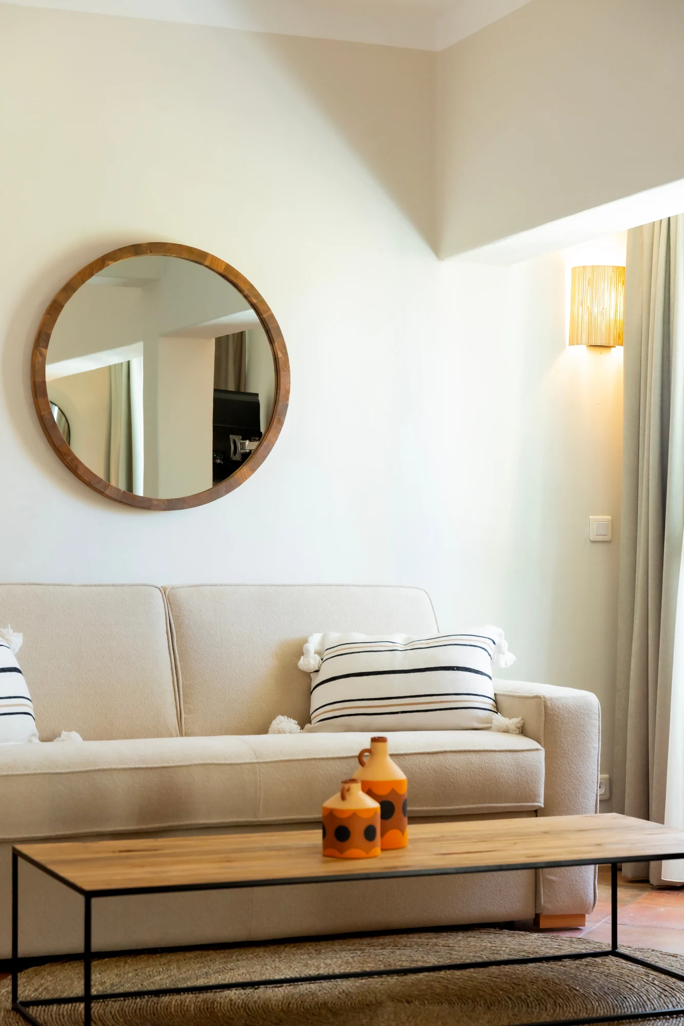 Salon lumineux avec canapé beige orné de coussins blancs à rayures noires, table basse en bois avec pieds en métal noir, deux vases décoratifs orange et noir, miroir rond encadré de bois, et lampe murale jaune près d'une fenêtre avec rideaux clairs.