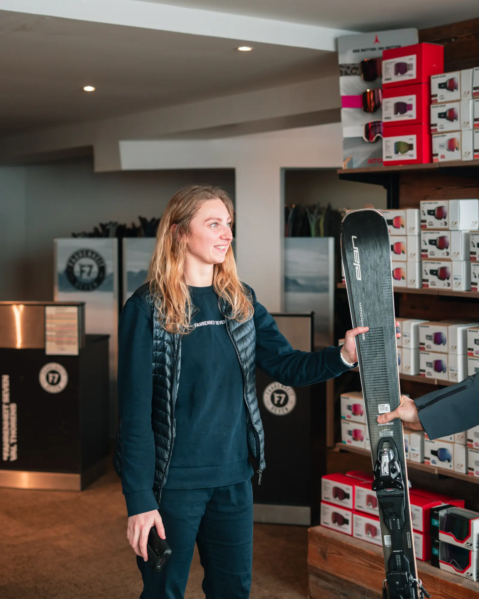 Une femme souriante en tenue décontractée tenant un ski Elan, reçevant le ski d'une autre personne dans un magasin de sports d'hiver avec des boîtes de lunettes de protection en arrière-plan.