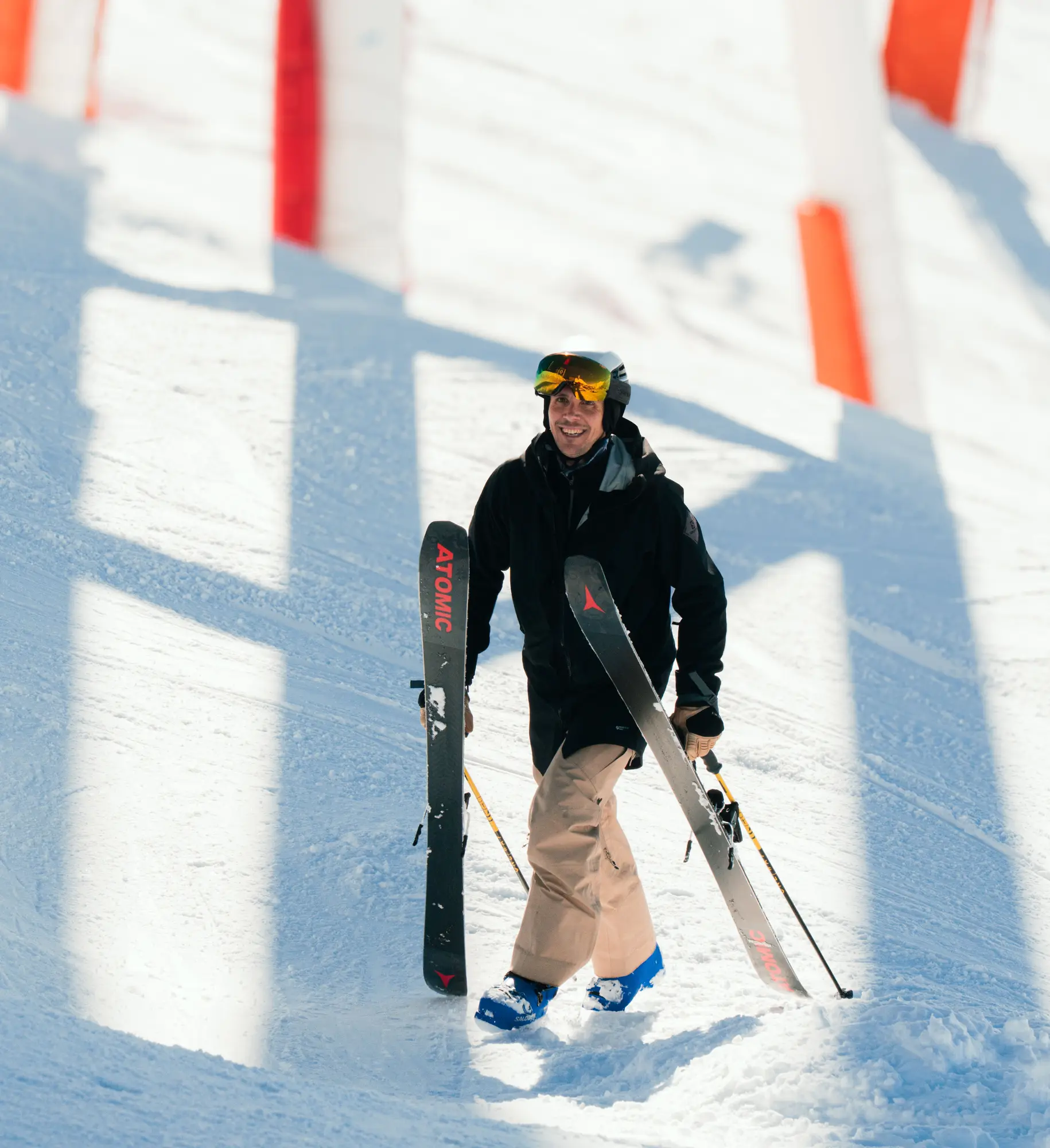 Homme souriant debout dans la neige portant une veste noire, un pantalon beige, des bottes bleues et un casque avec lunettes de ski, tenant des skis Atomic et des bâtons de ski.