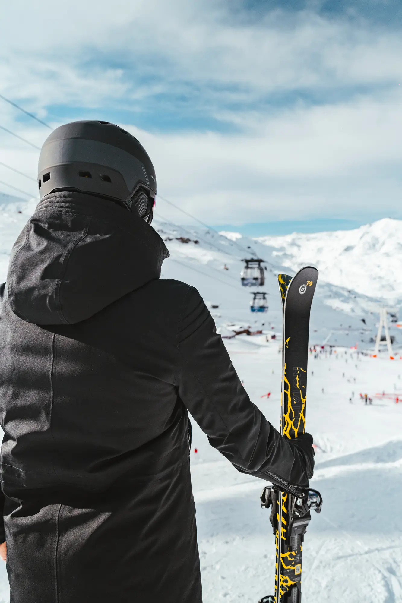 Personne vue de dos portant un casque et une veste noire, tenant des skis noirs et jaunes avec des montagnes enneigées et une station de ski en arrière-plan.