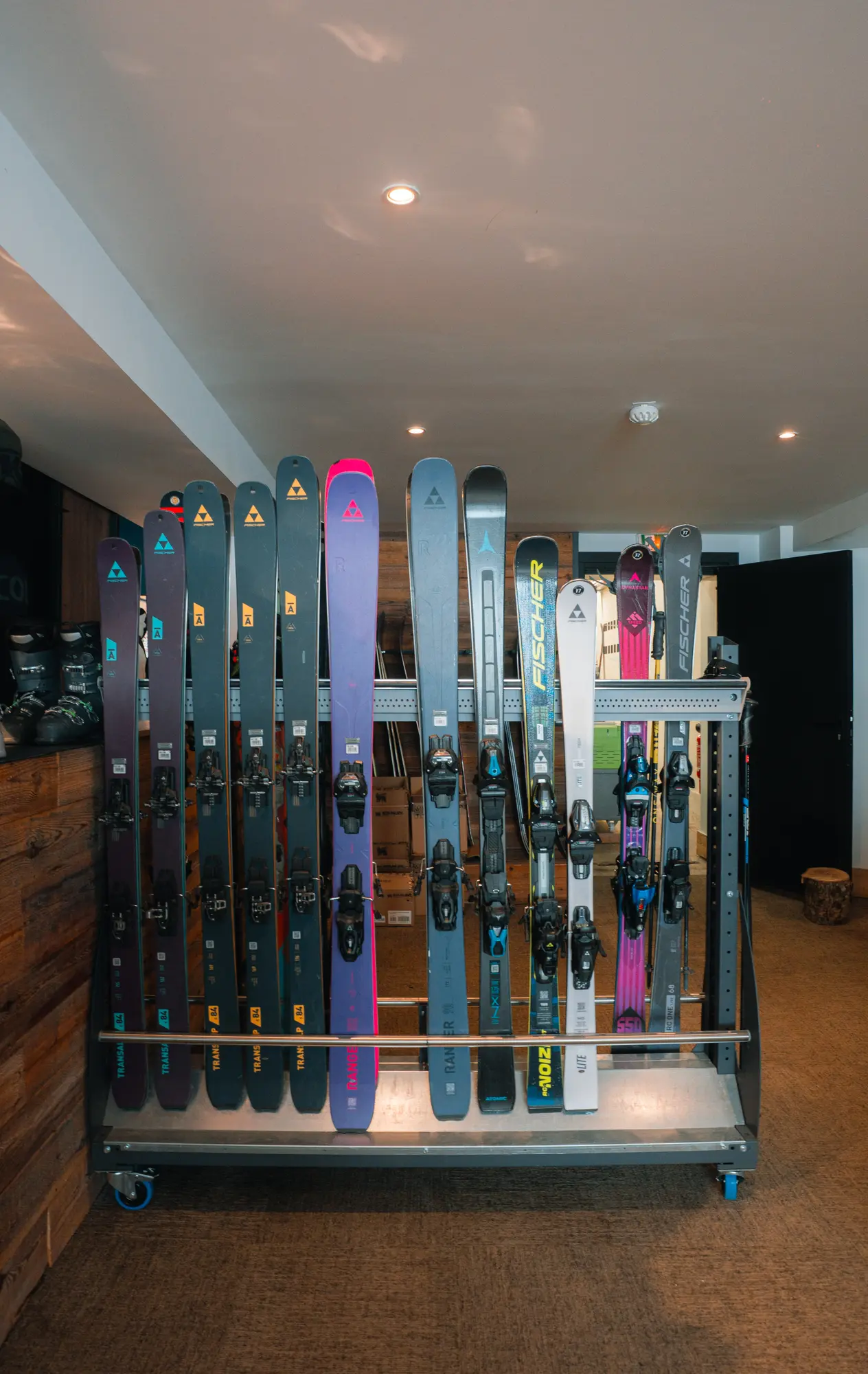 Un support métallique contenant plusieurs paires de skis alignés verticalement dans une pièce intérieure. Le sol est recouvert de moquette et les murs sont en bois et blanc. Le support est équipé de roulettes bleues et il y a des chaussures de ski posées à gauche sur une étagère en bois.