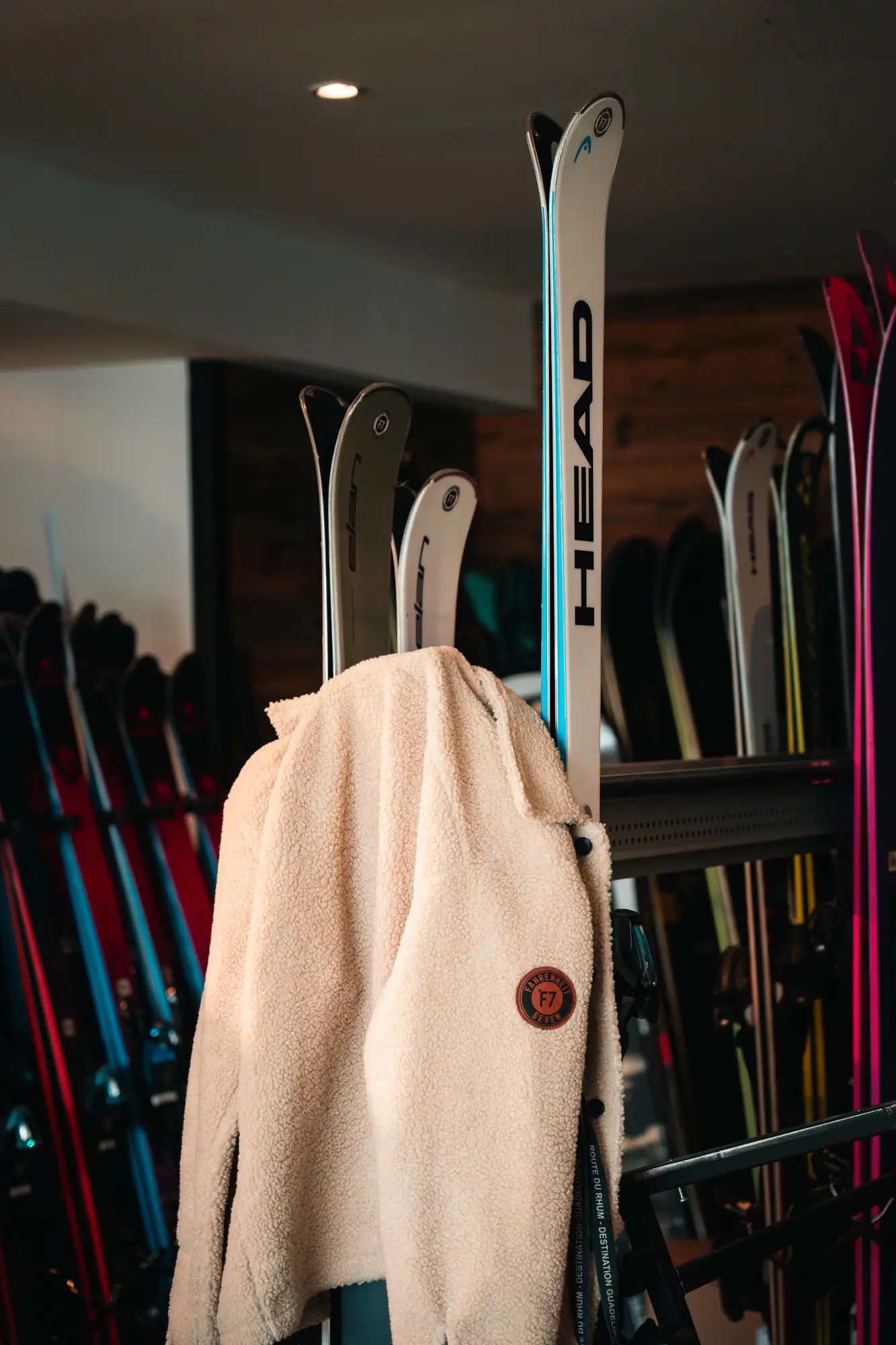 Veste en laine beige suspendue sur une paire de skis HEAD dans un local avec plusieurs paires de skis rangées en arrière-plan.