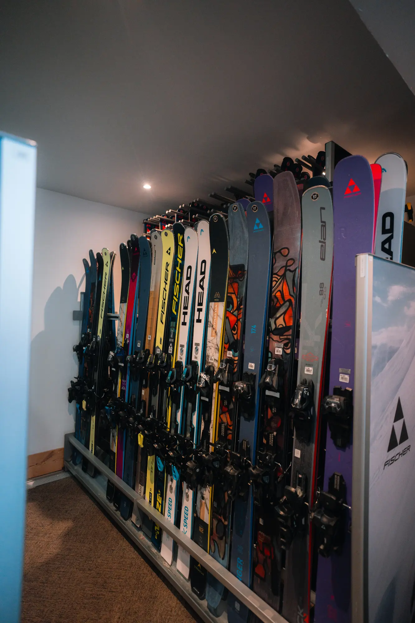 Rangée de skis colorés alignés verticalement dans un casier de rangement intérieur avec divers modèles et marques visibles.