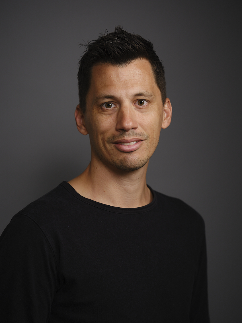 Un homme aux cheveux courts et foncés porte un t-shirt noir et sourit légèrement devant un fond gris uni.