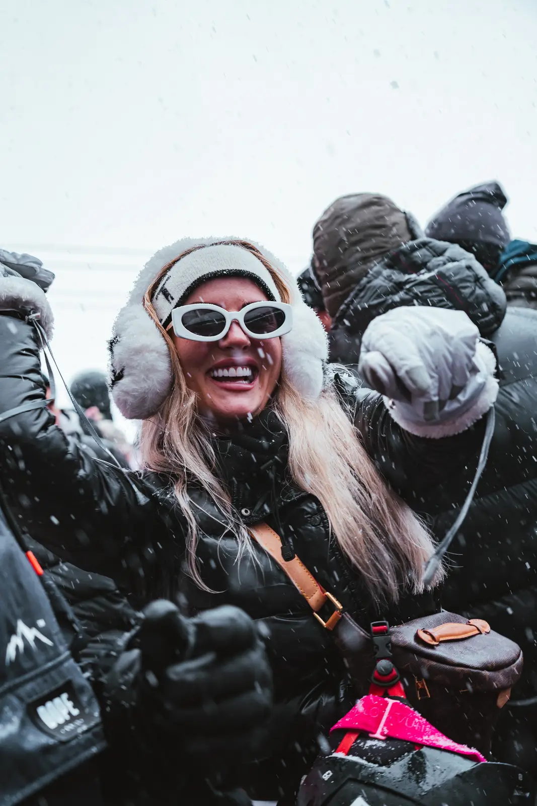 Jeune femme souriante portant des lunettes de soleil blanches, un bandeau et des cache-oreilles en fourrure, gantée, et vêtue d'une veste noire, célèbre sous la neige dans une foule habillée chaudement.