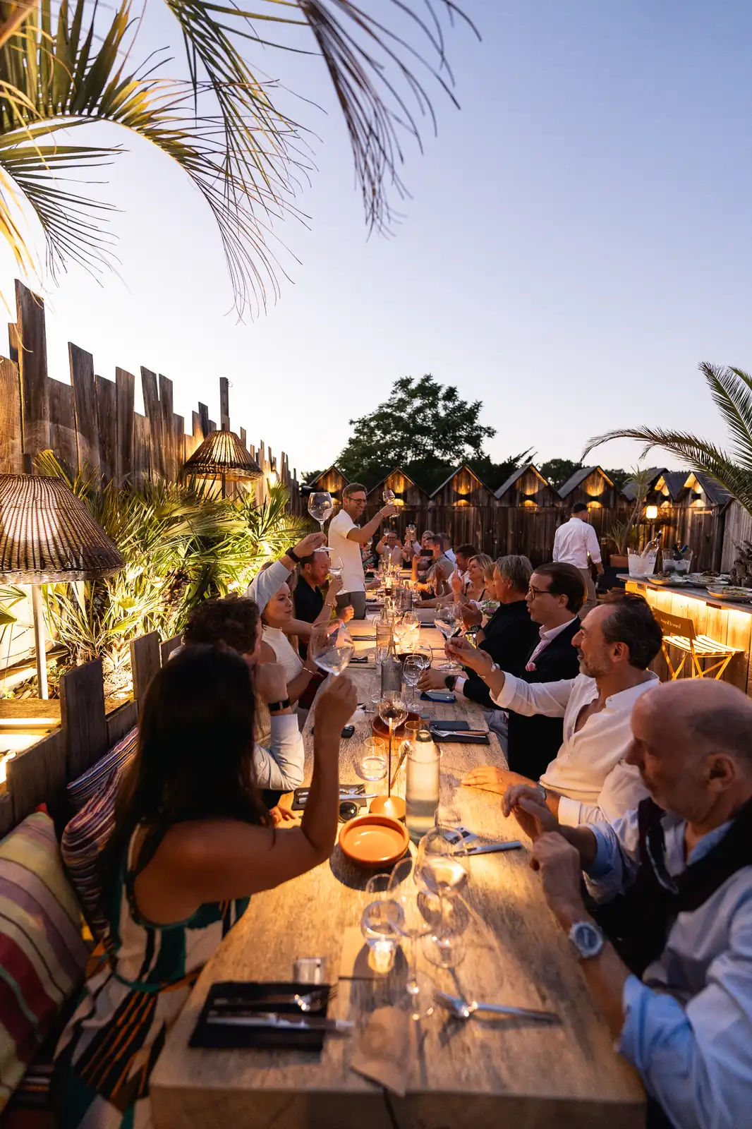 Groupe d'adultes assis à une longue table en bois en extérieur au crépuscule, levant leurs verres pour un toast, entourés de plantes et de lumières chaleureuses dans une ambiance conviviale.