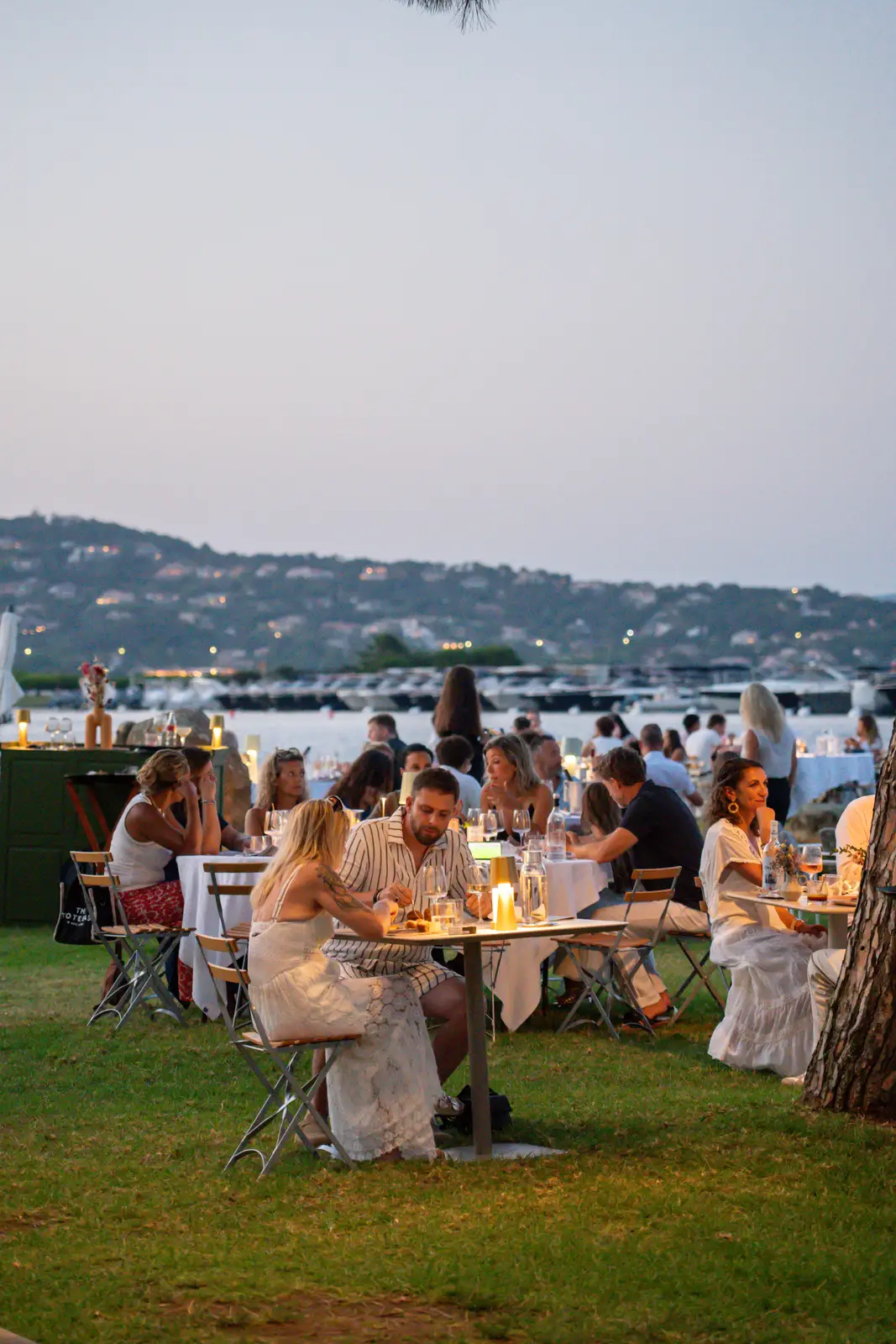 Des personnes dînent dehors en soirée sur des tables avec nappes blanches, avec vue sur un port rempli de bateaux et des collines en arrière-plan.