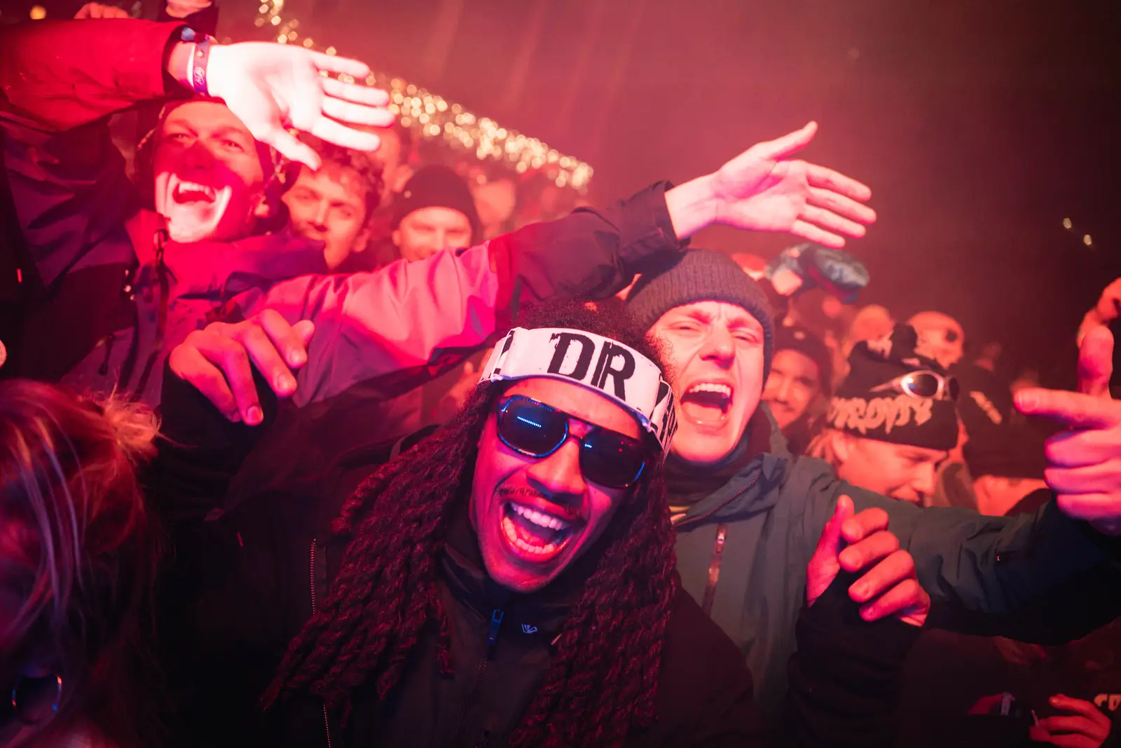 Un groupe de personnes souriantes et enthousiastes lors d'un événement en soirée, avec un homme portant des lunettes de soleil et un bandeau blanc inscrit DR au centre.
