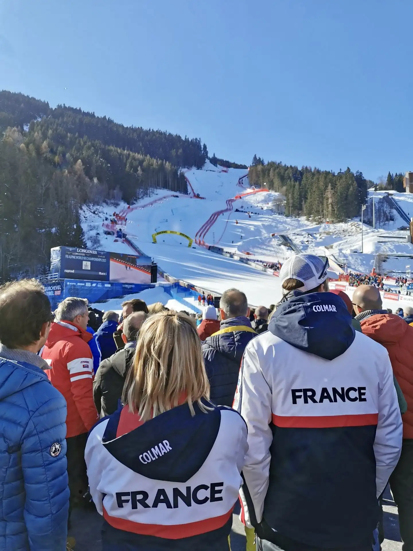 Spectateurs portant des vestes blanches, bleues et rouges avec l'inscription FRANCE regardant une piste de ski enneigée avec des barrières rouges lors d'une compétition, sous un ciel bleu clair et entourée de forêts.