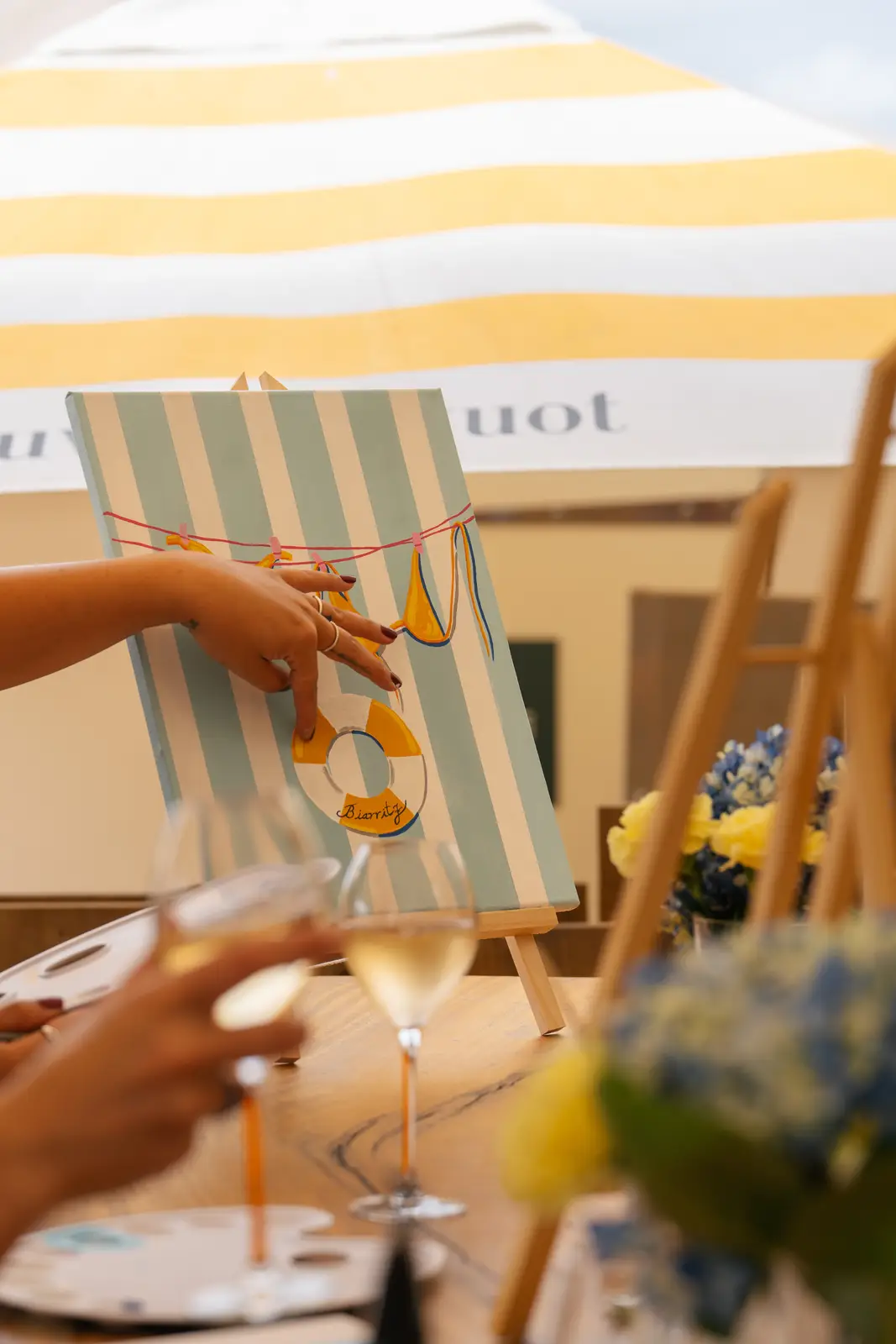 Une personne peignant sur une toile à rayures bleu clair et blanc, représentant un maillot de bain et une bouée jaune accrochée à une corde. Deux verres de vin blanc sont posés sur la table en bois devant la toile, avec des fleurs jaunes et bleues en arrière-plan, sous un parasol rayé jaune et blanc.