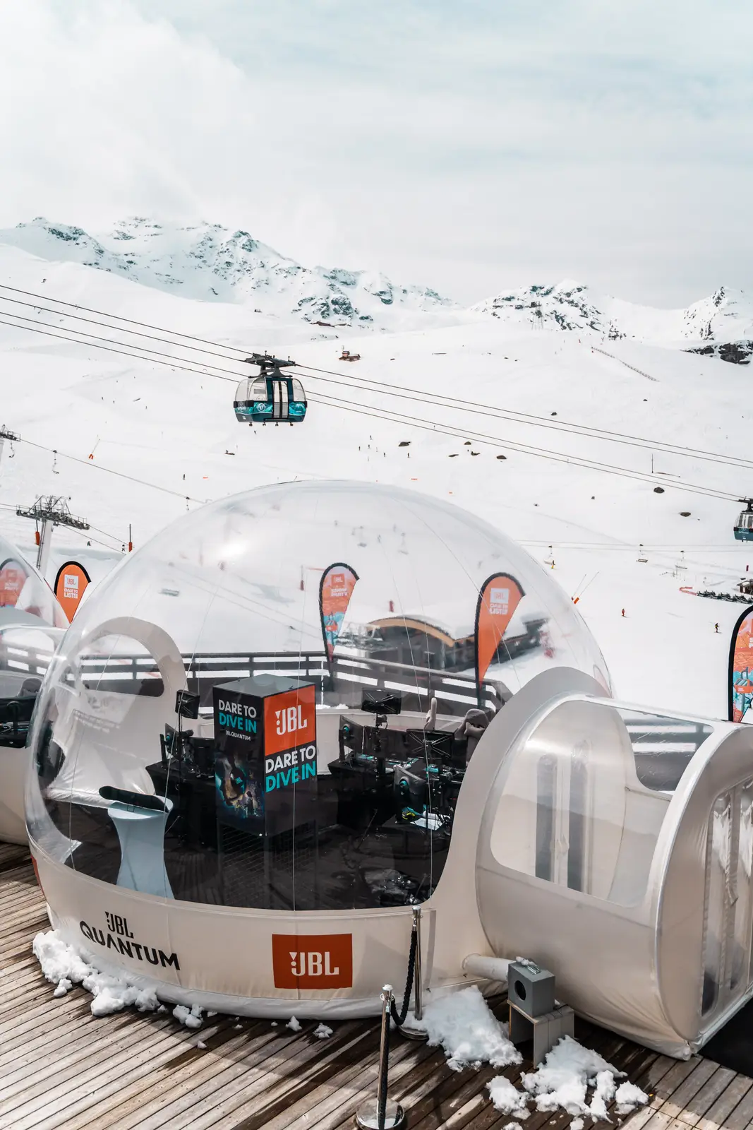 Dôme transparent JBL Quantum installé sur une terrasse en bois enneigée dans une station de ski avec des montagnes enneigées et des télécabines en arrière-plan.