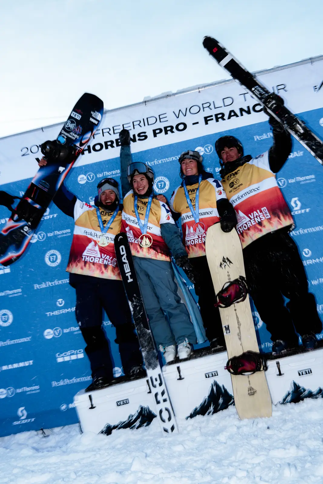 Quatre athlètes de sports d'hiver portant des médailles et des vestes jaunes avec des logos, debout sur un podium enneigé lors du Freeride World Tour à Val Thorens, France, tenant un snowboard et des skis, célébrant leur victoire.