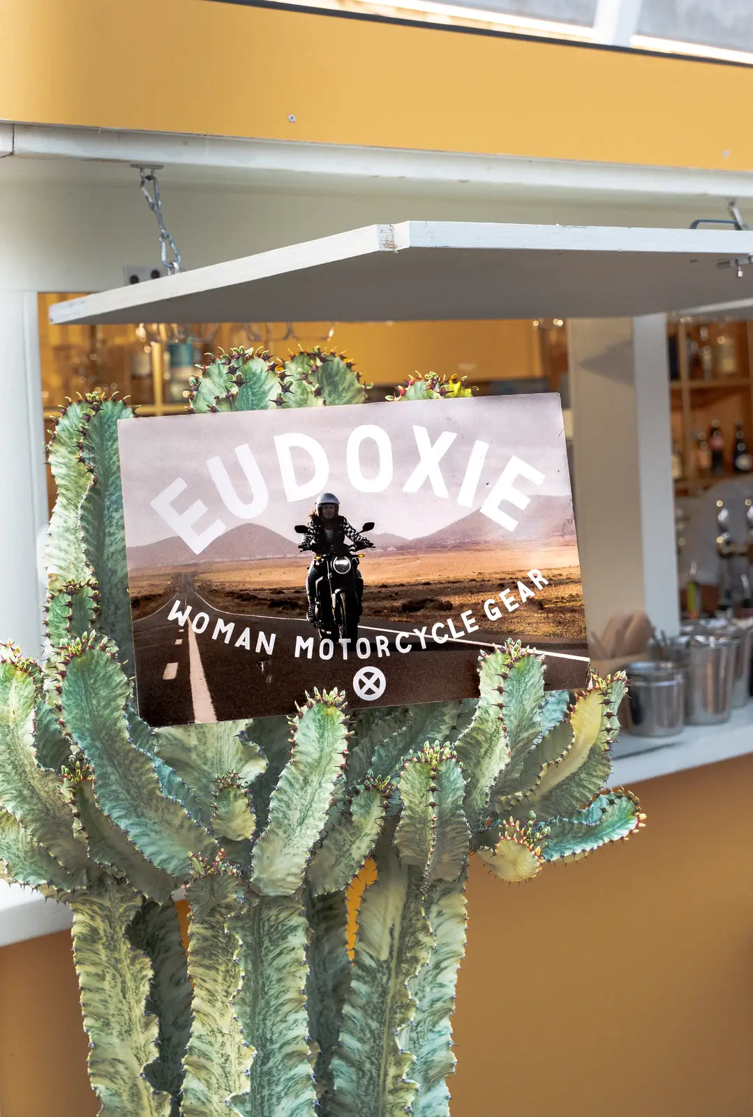 Affiche suspendue devant un cactus montrant une femme en moto sur une route désertique avec le texte « EUDOXIE WOMAN MOTORCYCLE GEAR ».