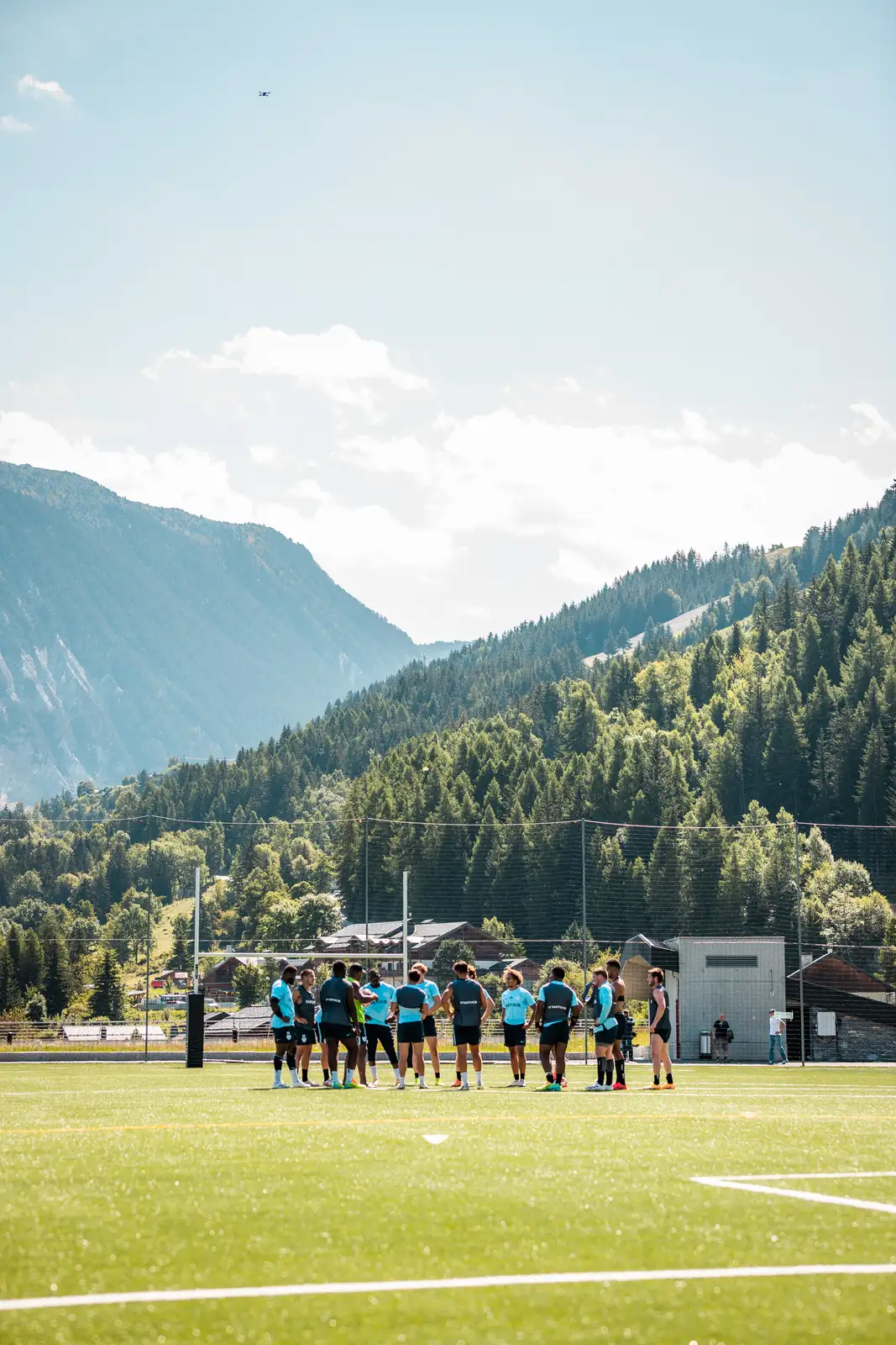 Une équipe de joueurs de rugby en tenue d'entraînement bleu clair et noir est rassemblée au centre d'un terrain vert entouré de montagnes boisées sous un ciel clair.