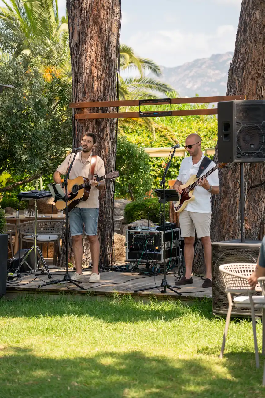 Deux musiciens jouent sur une petite scène en plein air devant de grands arbres et une végétation luxuriante, l'un chantant en jouant de la guitare acoustique, l'autre jouant de la guitare basse électrique, avec des équipements audio autour d'eux et une chaise en osier au premier plan.