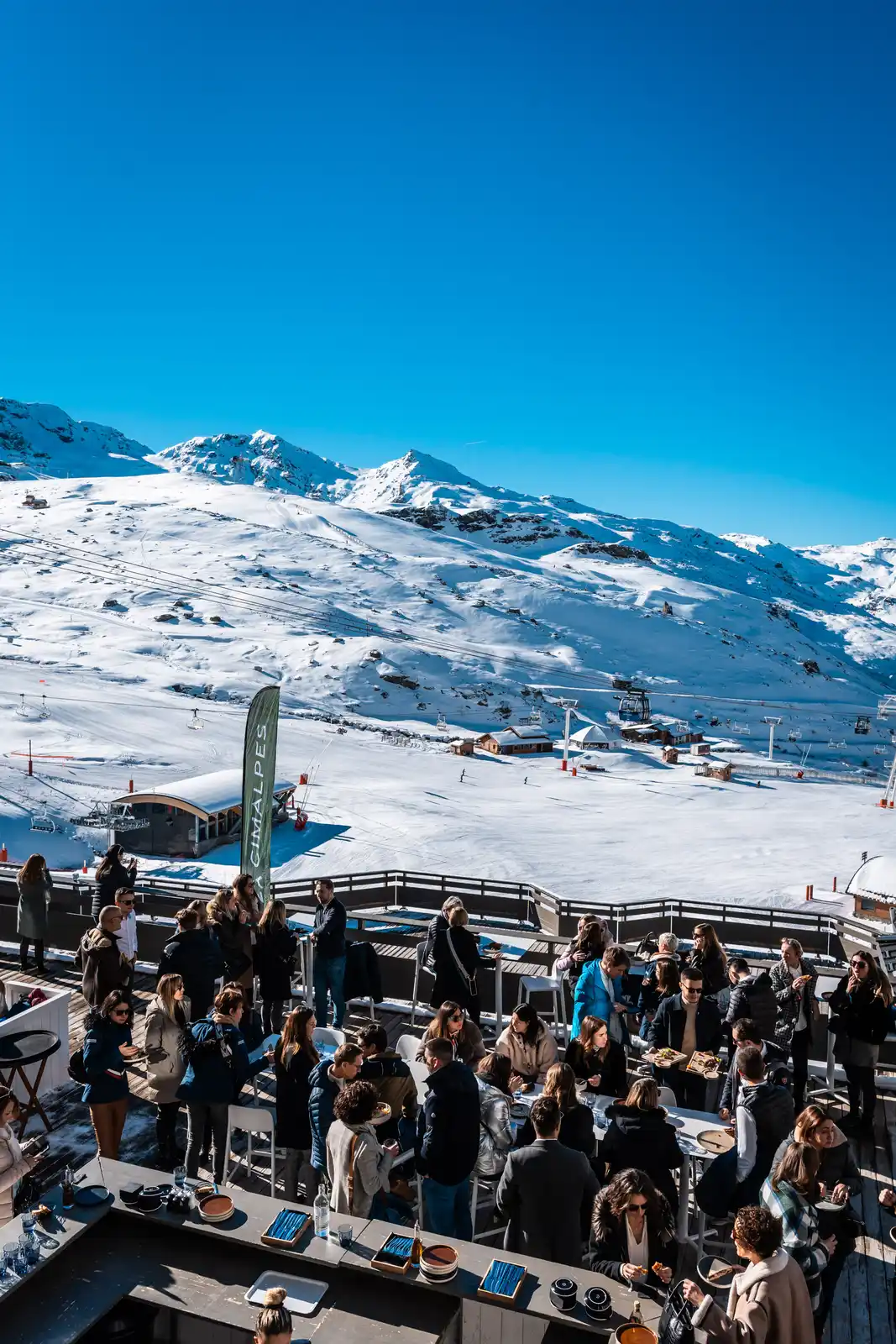Groupe de personnes réunies sur une terrasse en bois ensoleillée avec vue sur une station de ski enneigée et des montagnes en arrière-plan sous un ciel bleu clair.