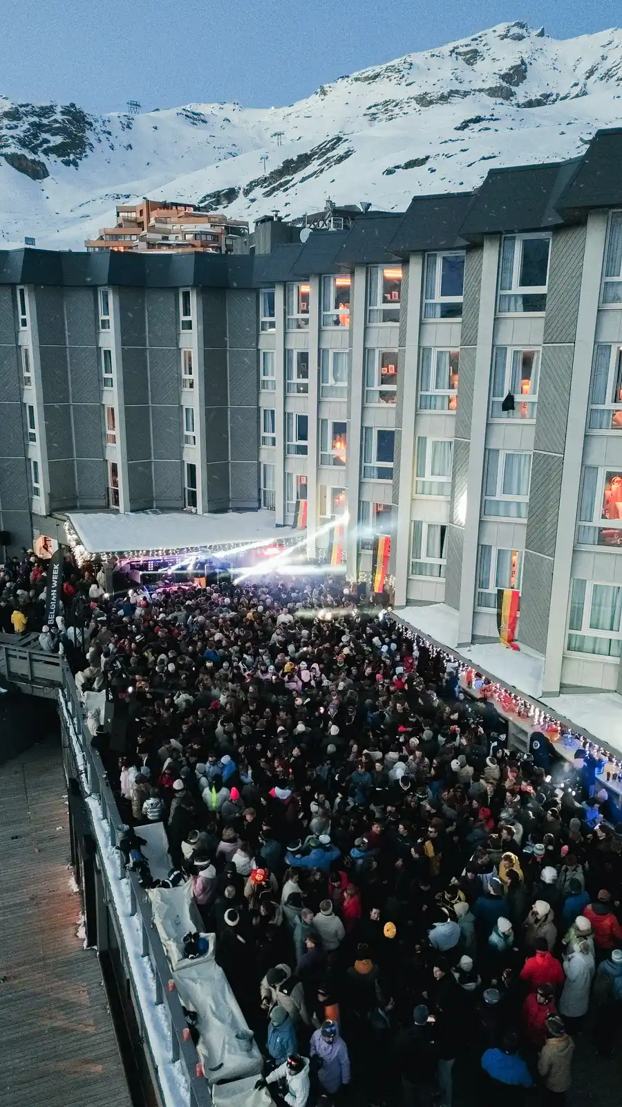 Foule nombreuse regroupée devant un bâtiment moderne avec plusieurs étages, décoré de drapeaux belge et lumières festives, en montagne enneigée au crépuscule.